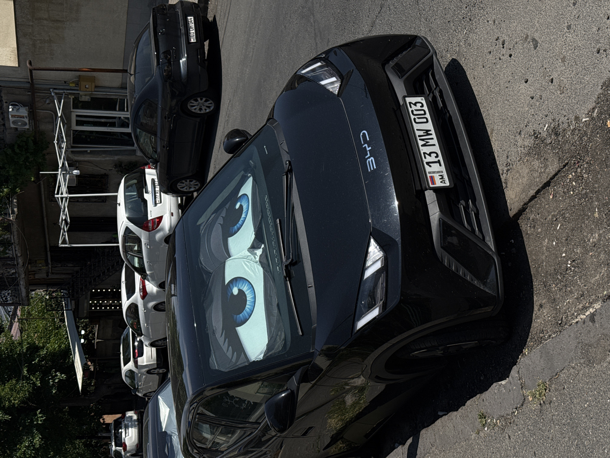 A black BYD electric car, adorned with sunshades shaped like large blue eyes on the windshield, is parked on a city street. The car has an Armenian license plate reading "13 MW 003" with the Armenian flag emblem. In the background, other cars, including white sedans and SUVs, are parked along the street, and a building with an air conditioning unit and an outdoor awning is visible. The scene is bathed in daylight, casting shadows on the pavement.