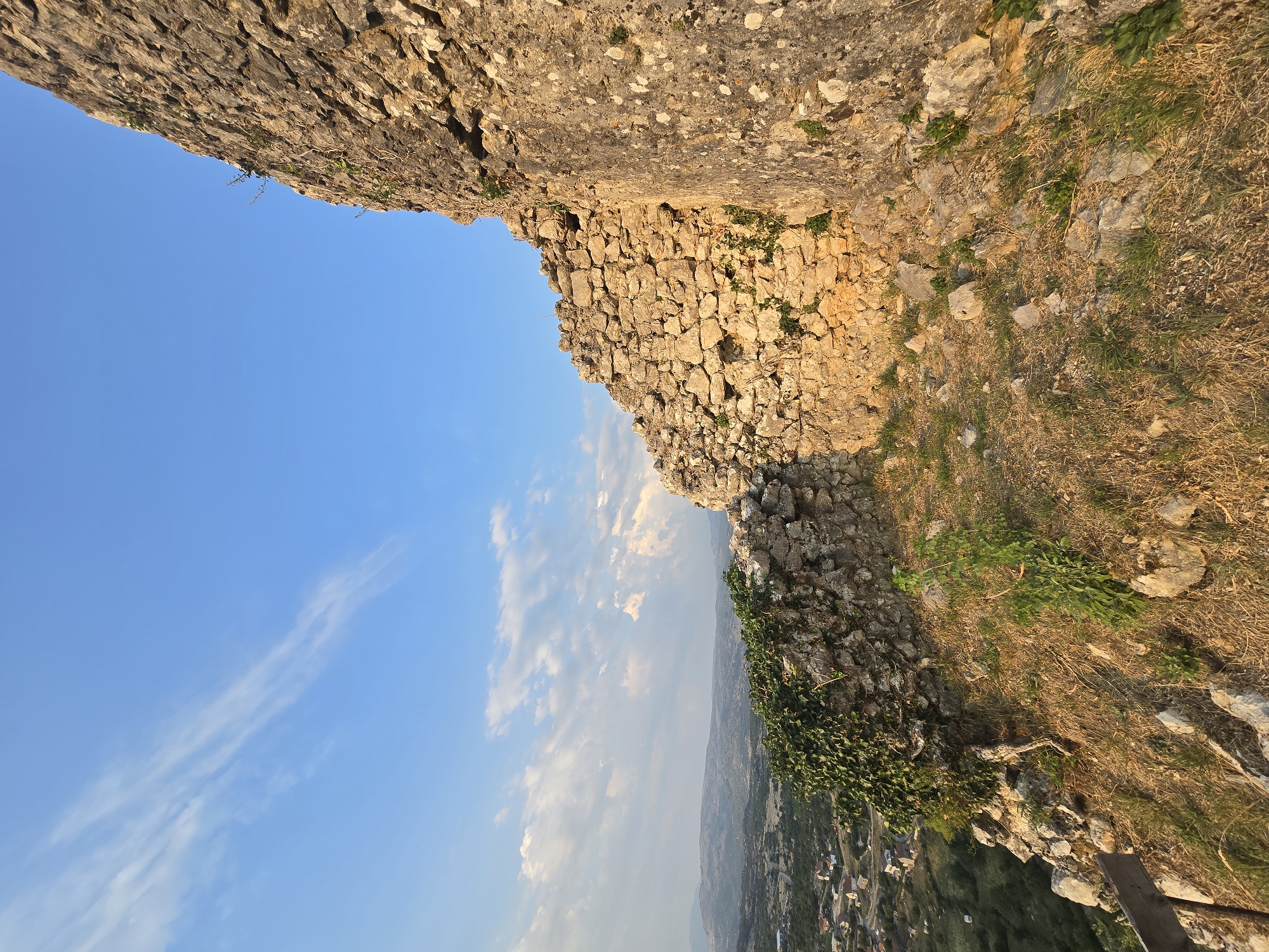 The image depicts a view from within the ruins of Medun fortress, located in the city of Medun, Montenegro. The foreground is dominated by a rough, stone wall, partially overgrown with green ivy on the left side. The wall is constructed from irregular, light-colored stones of varying sizes. Dry, yellow grass and scattered rocks cover the uneven ground in front of the wall.
In the middle ground, the remains of the fortress wall continue, curving away to the left. Beyond the ruins, a landscape unfolds with rolling hills covered in green vegetation and dotted with small buildings, suggesting a rural or semi-urban setting. Further in the distance, a hazy, mountainous terrain is visible under a clear blue sky.
The sky occupies the upper portion of the image, with a few wispy white clouds scattered across it. The sunlight, likely from a setting sun, casts a warm, golden glow on the stone structures, highlighting their texture and creating long shadows. The overall scene conveys a sense of ancient history and natural beauty, viewed from a high vantage point overlooking the surrounding area. There are no people or animals visible in the image.