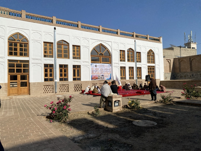 A two-story building with arched windows and a decorative facade serves as a backdrop for an outdoor gathering. People sit on a red rug. A banner hangs on the building. A microphone and stand face the group. A flower garden borders the brick-paved area. Another building is visible in the background. The sky is clear and bright.