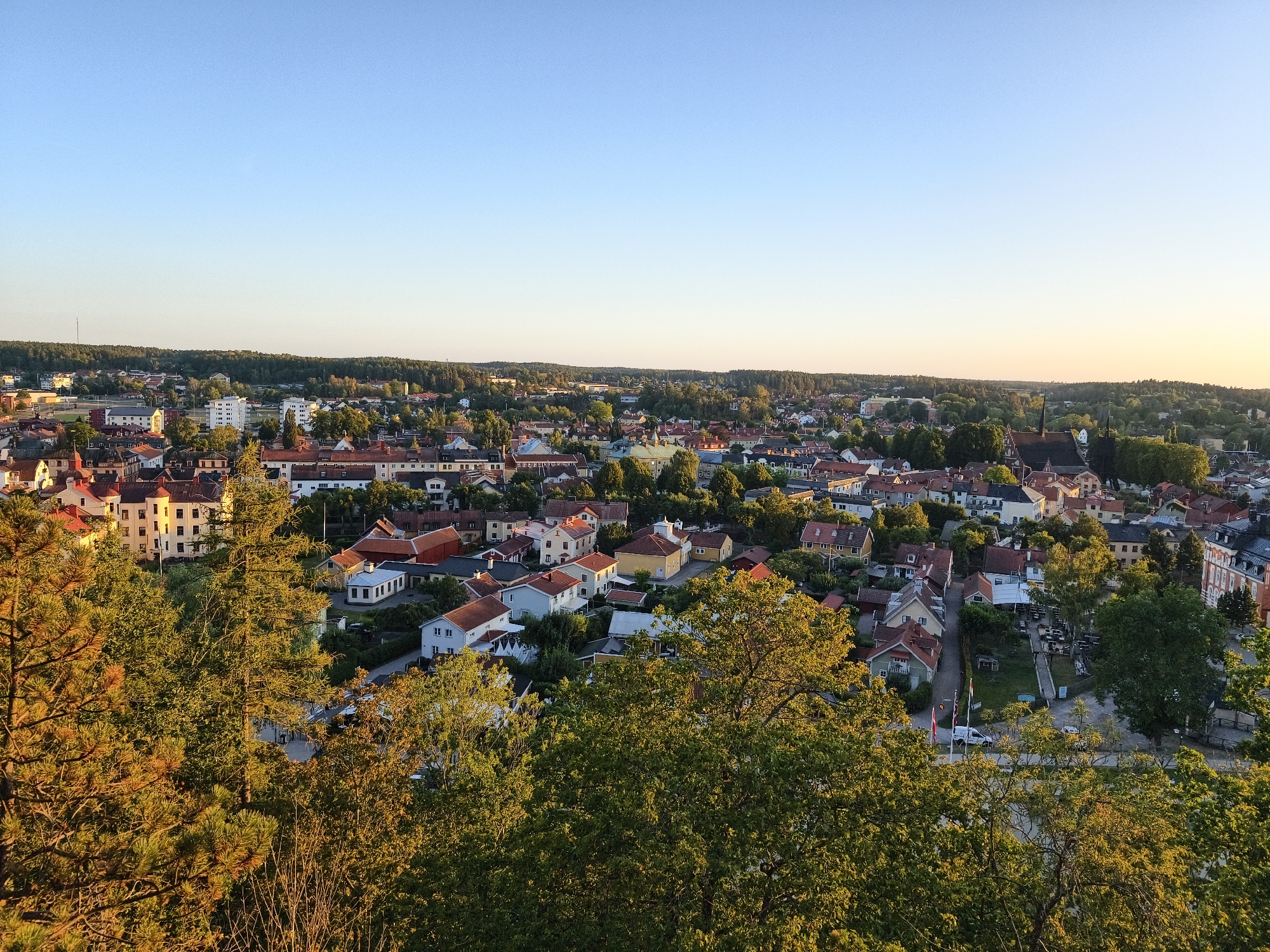 This is a high-angle, wide shot of the town of Söderköping, Sweden, taken during the golden hour. In the foreground, lush green trees with hints of autumn gold are visible, obscuring part of the view. Beyond the trees, a dense residential area unfolds, characterized by a mix of single-family homes and multi-story buildings with red-tiled and dark-colored roofs. The architecture suggests a historical European setting, with some buildings featuring light-colored facades and red-brick accents. A prominent church with a tall, dark spire stands out in the mid-ground. The town is nestled amongst rolling hills covered in dense forest, extending to the horizon. The sky is a clear, pale blue, transitioning to a warmer, orange hue near the horizon, indicating either sunrise or sunset. There are no visible people in the image. The overall impression is one of a peaceful, established European town surrounded by natural beauty.