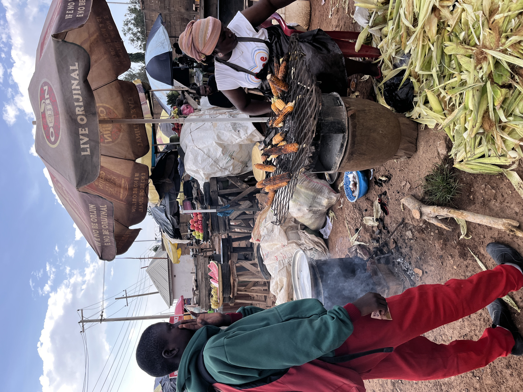 This image captures a vibrant outdoor market scene in Gura Top, Nigeria, under a bright, partly cloudy sky. The central subject is a woman, wearing a light-colored headwrap and a white t-shirt, actively grilling corn cobs on a round, elevated charcoal grill. Several corn cobs, some appearing well-roasted, are arranged on the grill. Adjacent to her stall is a substantial pile of raw corn cobs and husks.
A large brown market umbrella dominates the left side of the frame, providing shade and bearing prominent branding: "ORIJIN", "LIVE ORIJINAL", and the Yoruba phrase "IFE NO BE ORIJIN ENO BE ORIJINAL" (If it's not Orijin, it's not original). Another umbrella, black and white, is visible in the background. Behind the woman, market stalls are set up with wooden frames, displaying various goods including large white sacks and fresh produce like red apples, bananas, and a slice of watermelon.
In the foreground, a person dressed in a green hoodie, red top, and red pants stands, looking towards the sky. A small fire pit with rising smoke is on the dusty ground near their feet. The overall setting is unpaved and features utility poles with power lines and glimpses of distant buildings under the clear sky. The word "VICE" is also partially visible on a sign in the background. The scene depicts a typical street food vendor in an active, bustling market.