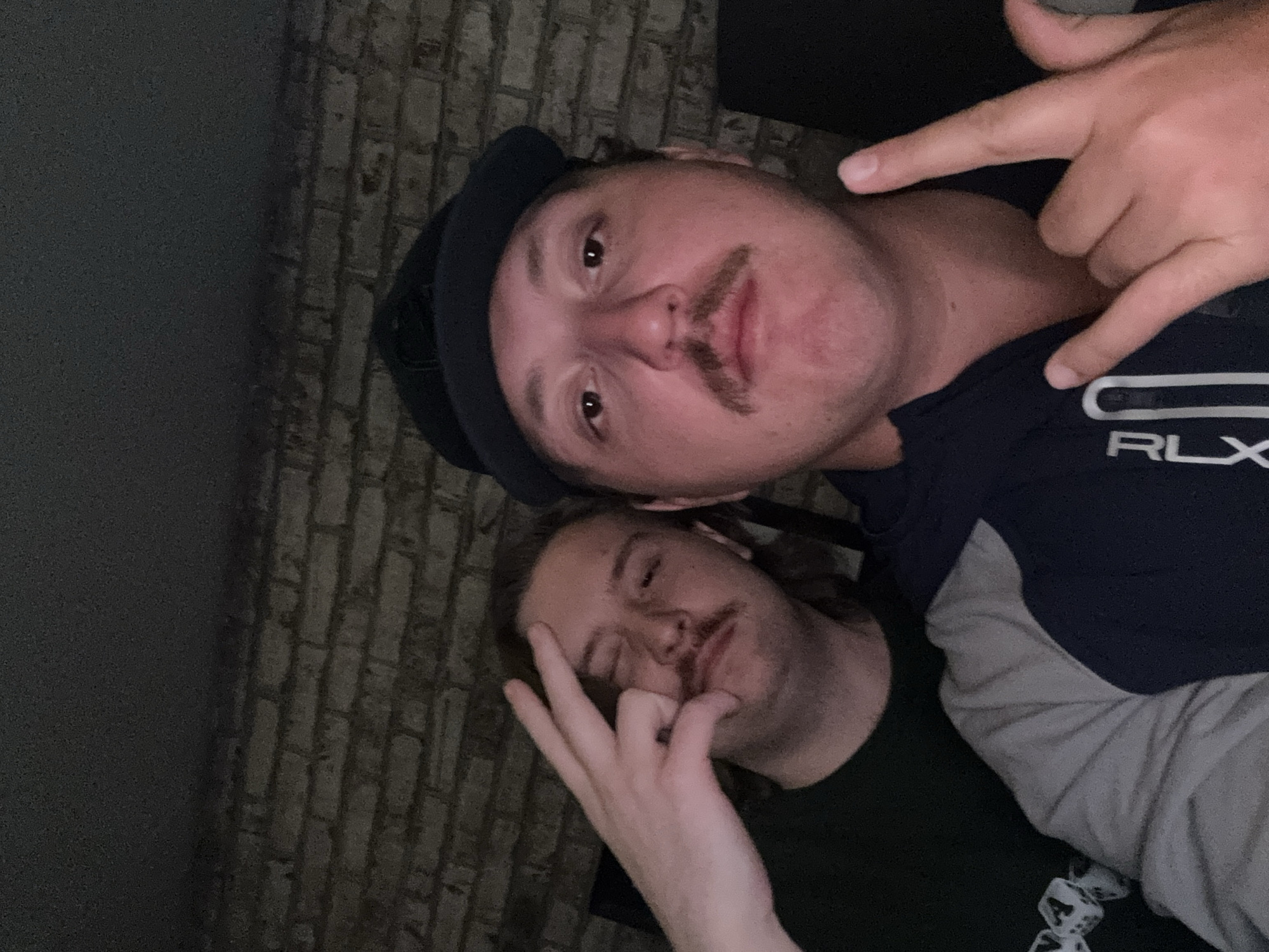 Two males are posing for a selfie-style photograph indoors near a brick wall. The male in the foreground is wearing a navy cap and a navy and grey RLX branded shirt. He has brown hair and a mustache, and he is making a hand gesture with his right hand. The male in the background also has brown hair and a mustache, and he is wearing a dark-colored shirt and making a hand gesture. The photo appears to have been taken in dim lighting. There is no visible indication of the location being Huixquilucan de Degollado, Mexico, in the photo.