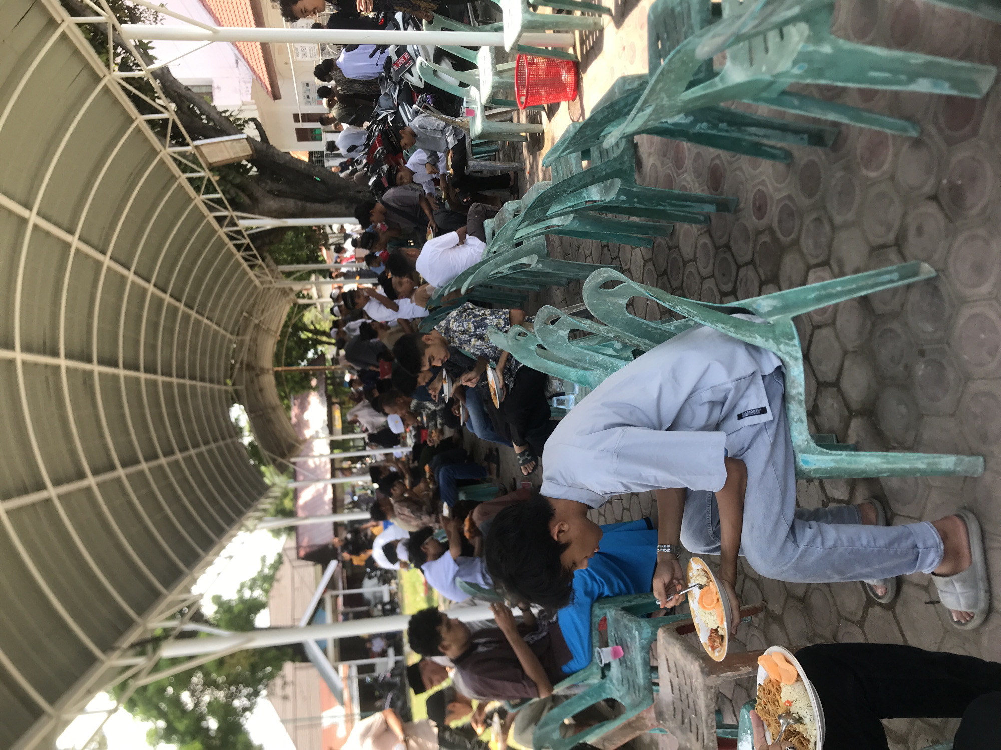 The image shows a large group of people seated on green plastic chairs under a metal-framed canopy. They appear to be eating from plates, with several individuals holding utensils. There is a red plastic basket visible in the background. Motorbikes are parked nearby. The scene is located in Lhokseumawe, Indonesia. The ground is paved with a hexagonal pattern.