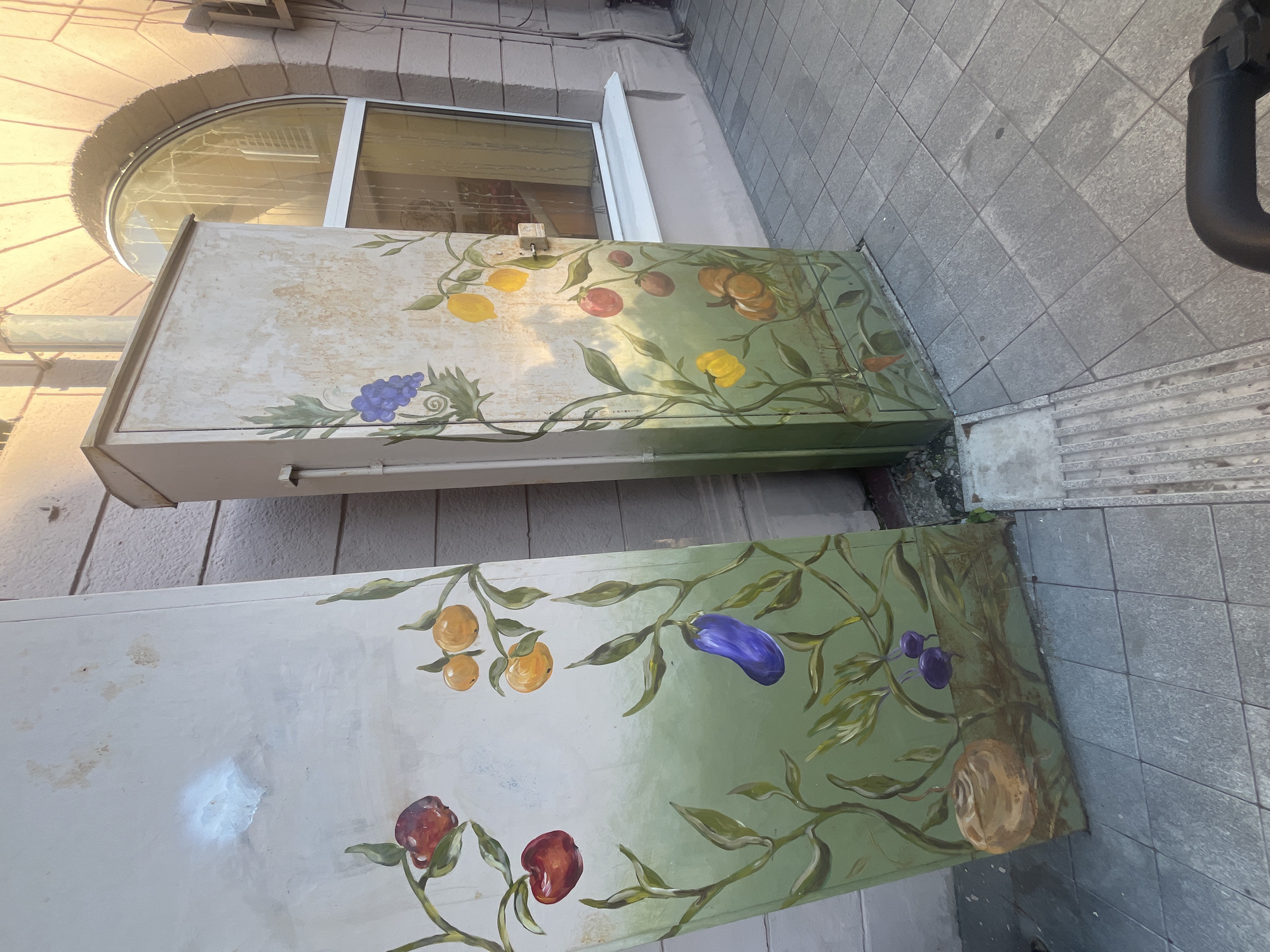 The image shows two large, rectangular objects, likely utility boxes, that have been painted with a fruit and vine motif. The objects are positioned outdoors on a paved surface, with gray square tiles forming the ground. To the right of the objects, a black pipe curves upwards, suggesting an industrial or urban setting.
The top object features a light-colored surface adorned with painted fruits including lemons, grapes, and what appear to be tomatoes or cherries. Green vines and leaves intertwine with the fruits, creating a decorative pattern that extends across the surface. The overall impression is one of an artistic embellishment on an otherwise utilitarian structure. The paint on this box shows some signs of wear and tear, with scuffs and a slightly faded appearance in places. Above and behind this object, a large, arched window is visible, set within a building with stone-like facade elements. The window reflects sunlight, indicating it is daytime.
The bottom object is similar in form but is painted with a predominantly green background that transitions into a lighter hue. This box also displays painted fruits and vines, with images of what look like oranges, an eggplant, and possibly plums or cherries. A circular, light-brown object, resembling a fruit or perhaps a decorative element, is painted on the right side of this box. The paint on this second box also shows some signs of wear, though it appears to be in slightly better condition than the top one.
The scene is likely set in a public outdoor space, possibly a street or courtyard. The sunlight suggests a clear or partly cloudy day. The decorative painting on the utility boxes adds an element of visual interest to the urban environment. There is no visible text in the image.