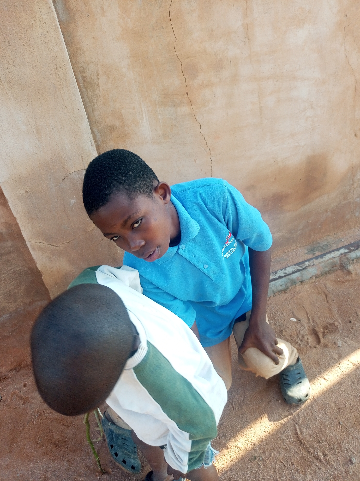 In Tempure, Nigeria, two young boys are seen in an outdoor setting. The boy in the foreground, viewed from behind, has dark, short hair and is wearing a white shirt with green and white stripes. He also wears dark clogs on his feet. The second boy, facing the camera, has dark hair and is wearing a bright blue polo shirt with a logo on the left chest. He is kneeling and appears to be adjusting or holding a light-colored object, possibly a piece of clothing or fabric, near his right leg, which is also clad in dark clogs. The ground beneath them is sandy and appears to be a dirt pathway. Behind the boys, a textured, light brown wall with visible cracks serves as the backdrop. Sunlight casts shadows on the ground, indicating it is daytime.