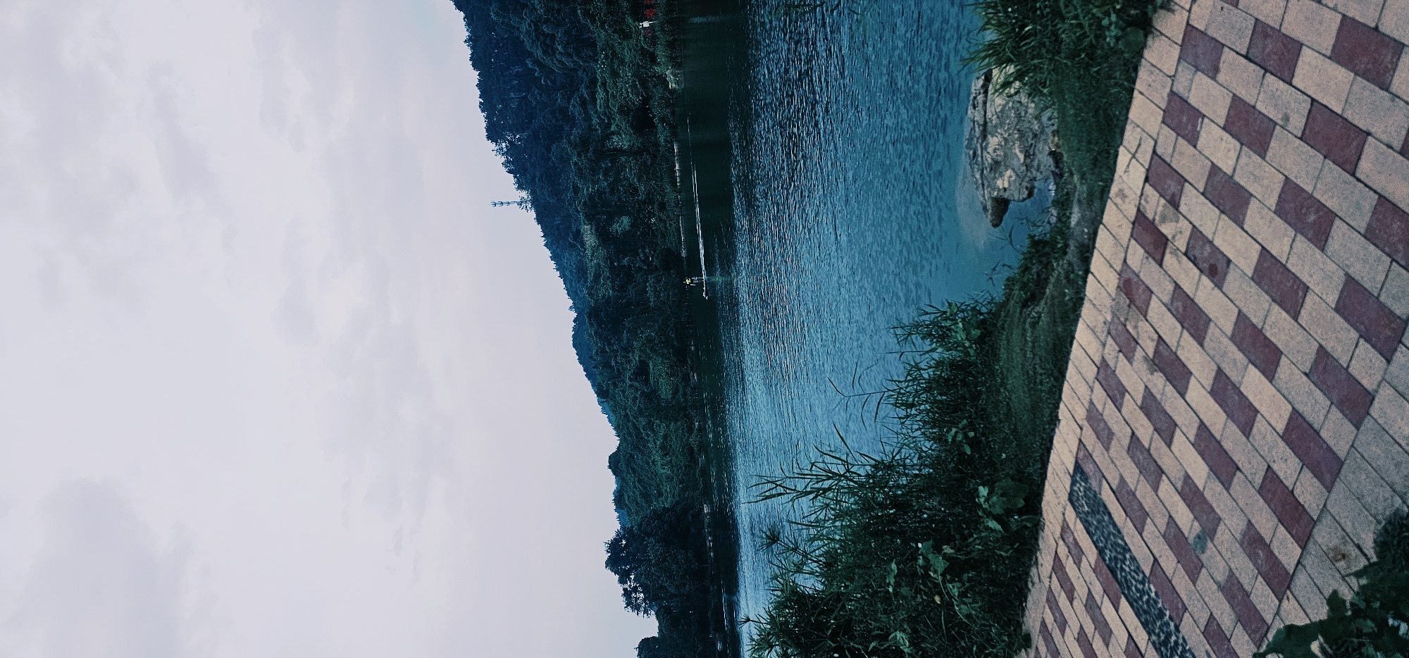 An overcast day at a body of water in Chongqing High-tech Zone, China. In the foreground, a paved walkway is visible, made of rectangular bricks in alternating shades of light tan and dark red, laid in a herringbone pattern. To the right of the walkway, a grassy embankment with tall reeds leads down to the water. A large, greyish rock sits on the edge of the embankment near the water.
The body of water, a lake or large pond, occupies the middle ground and stretches across the frame. Its surface is a deep blue-green with ripples, indicating a slight breeze. In the distance, a person on a paddleboard, wearing a yellow shirt, is visible on the water.
The background is dominated by a densely forested hillside that slopes upwards from the water's edge. The trees are a dark green, and the foliage appears lush. A white cellular tower is perched on the crest of the hill. The sky is a uniform pale grey, suggesting cloud cover. The overall impression is one of a serene, natural landscape within an urban or high-tech zone.