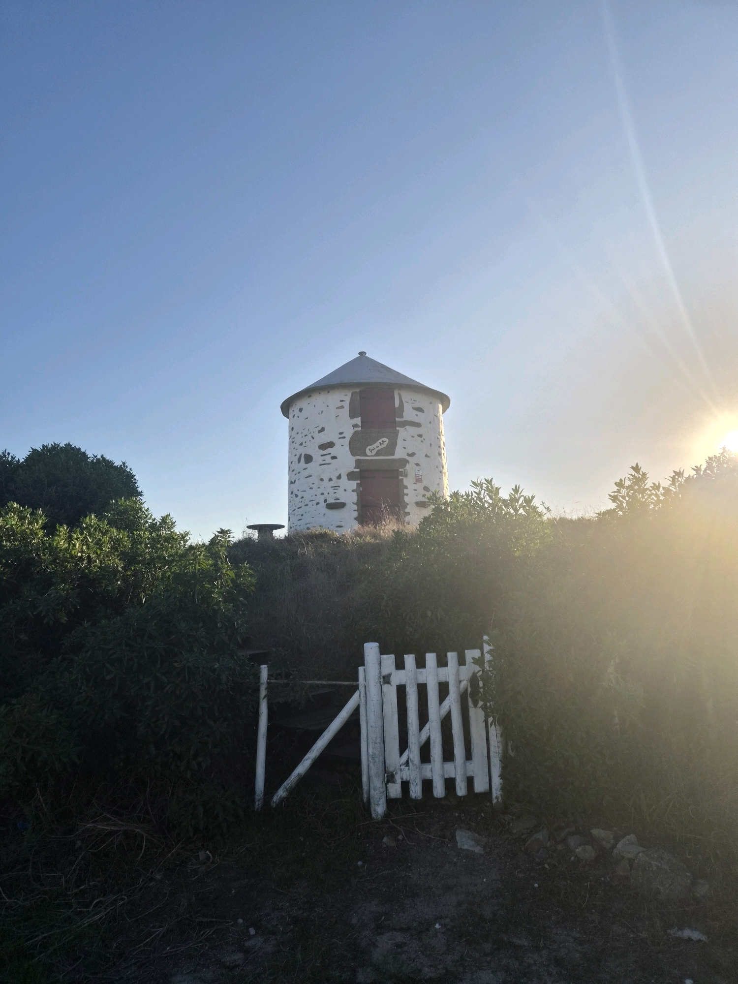 The image depicts a historic, whitewashed stone windmill with red doors, situated on a grassy hilltop. The windmill is partially obscured by lush green foliage and bushes, with a rustic white wooden gate in the foreground leading towards it. The sun is setting, casting a warm, golden glow over the scene and creating lens flare effects. The sky is a clear, bright blue, indicating fair weather. There is some text visible on the windmill, which appears to read "Feira da Noite". The overall mood is serene and picturesque, evoking a sense of timelessness and tranquility. The location context is Esposende, Portugal.