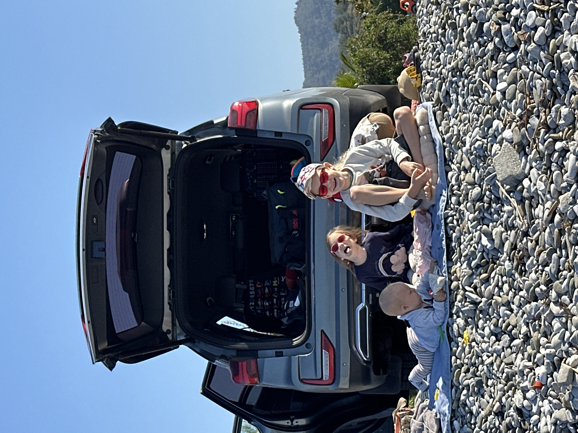 Three children and a silver car are on a pebble beach in Fethiye, Turkey. The car's back doors and trunk are open. Two girls sit on a blue cloth near the open trunk. A baby lies on the same cloth. The beach is covered in smooth, gray pebbles. In the background are trees and a mountain. The sky is blue.