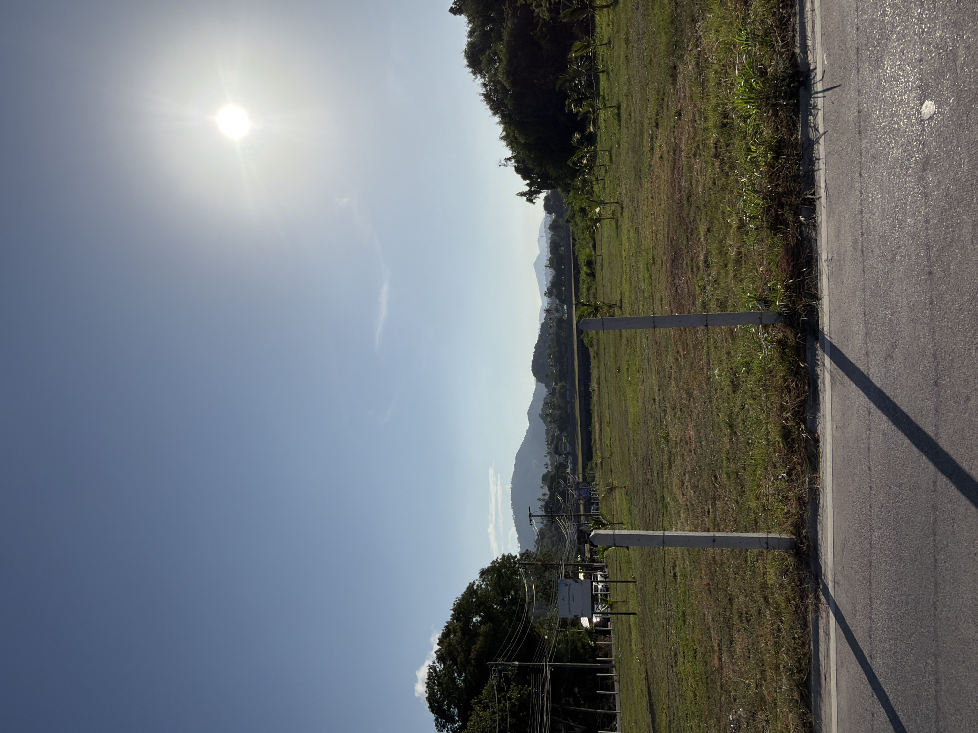 An image depicts a rural roadside scene in the Ko Samui District of Thailand. The foreground features an asphalt road with white lane markings and two long, dark shadows cast across it. Beyond the road, a sparsely vegetated field is visible, marked by a wire fence supported by two concrete posts. Several young trees are planted within the field. In the middle ground, a line of utility poles with numerous wires stretches horizontally, and beyond that, a cluster of buildings and more trees are present. The background reveals rolling, tree-covered hills under a bright, clear blue sky. The sun is prominently visible in the upper right quadrant of the sky, emitting a strong glare. The scene is devoid of any human presence.