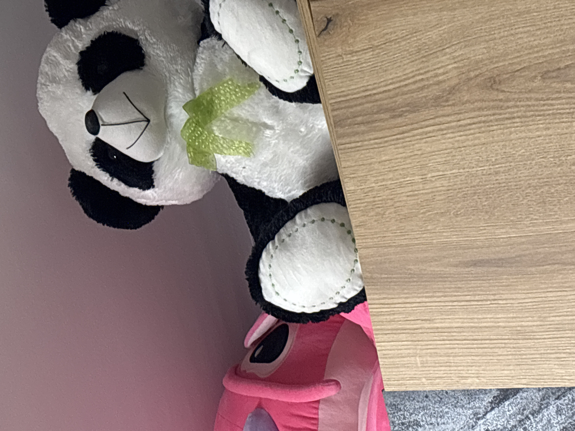 A close-up shot shows a stuffed panda bear and a pink stuffed animal situated partially under what appears to be a light brown wooden surface, likely a piece of furniture. The panda has a green bow. The environment includes a light pink wall and what appears to be gray carpet in the lower right corner. The scene does not provide enough information to determine the location in Burgas, Bulgaria.