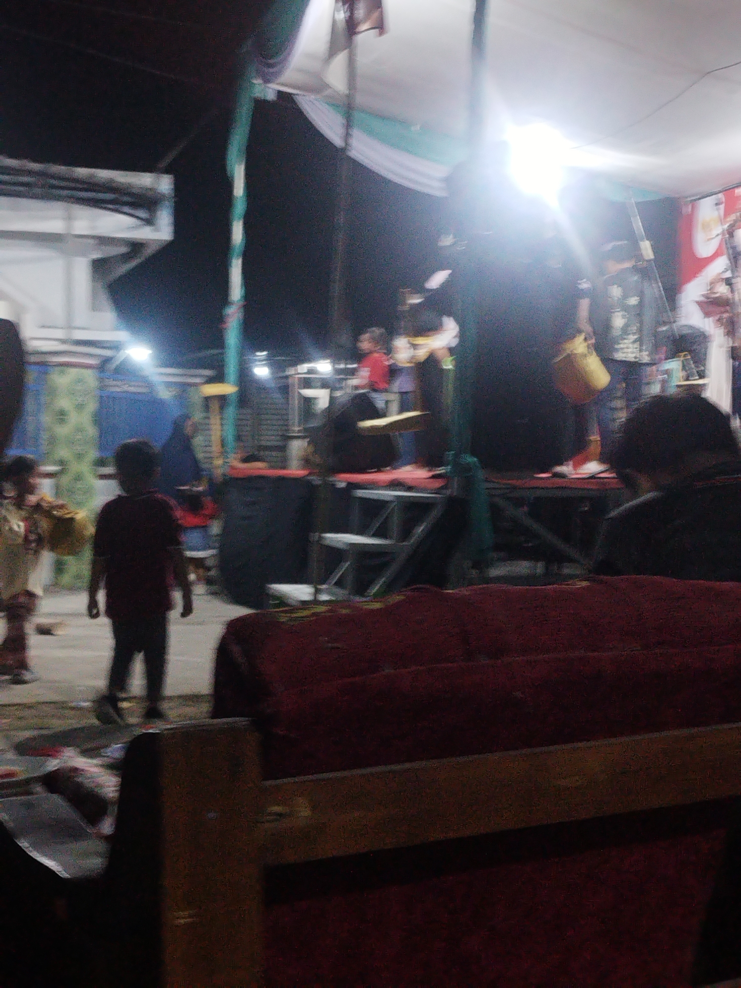 The image depicts an outdoor night scene, possibly at a community event or performance, in Kedungkumpul, Indonesia. In the foreground, a wooden bench with a dark red, plush surface is visible. Behind it, a stage area is set up, covered by a white tent with draped green and white fabric. Bright stage lights illuminate the area. On the stage, several individuals are present, with some appearing to be performers or stage crew. One person in the foreground on the left is wearing a maroon t-shirt and dark pants. To their left, another person, possibly a child, is wearing a patterned outfit and holding a yellow object. Further back, several other figures are seen on and around the stage. The background shows a dark sky and structures that suggest a village or town setting. The overall atmosphere appears to be festive and active, with a focus on the staged event.