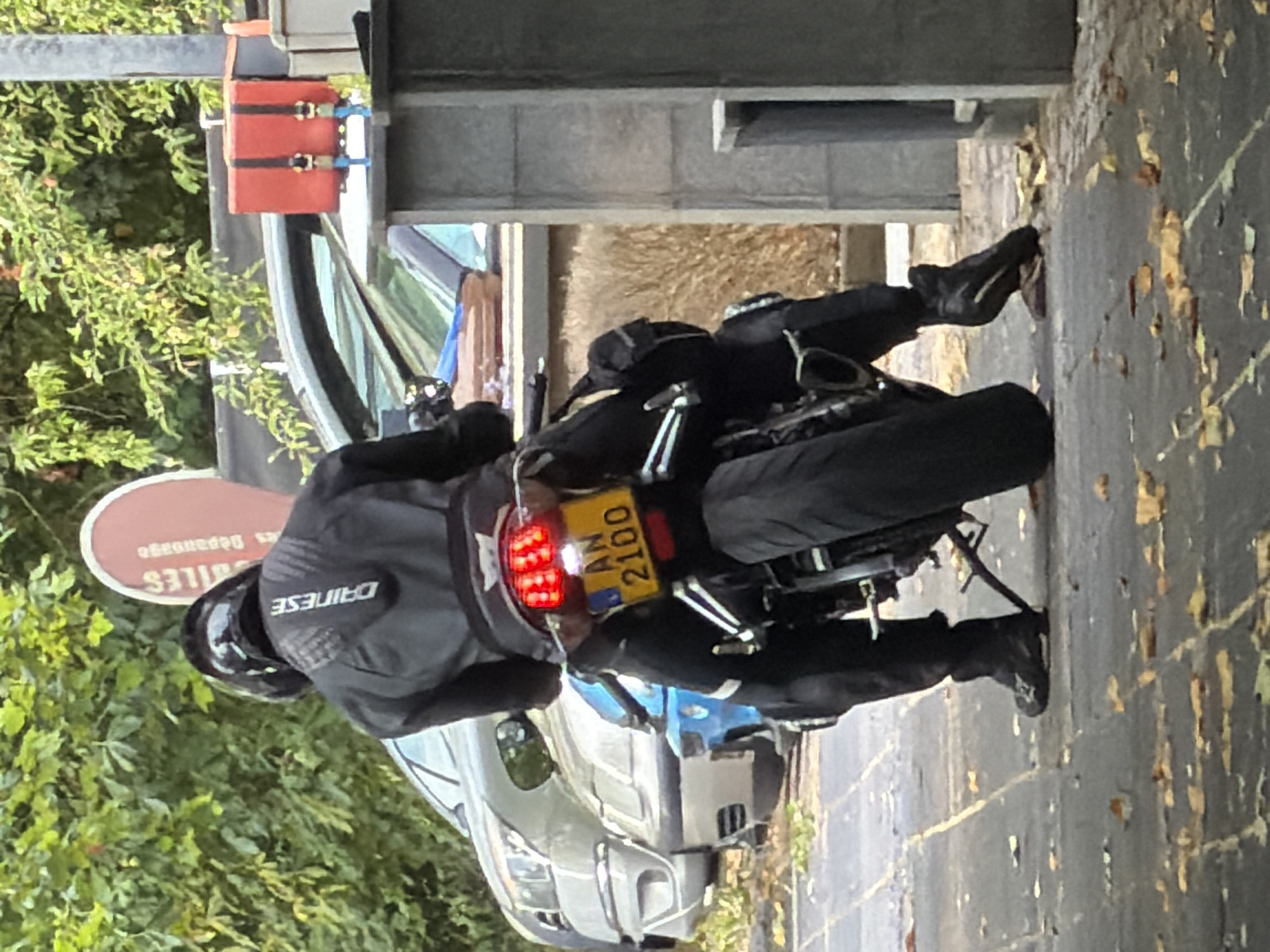 A person wearing a black motorcycle helmet and a black Dainese motorcycle jacket is seated on a motorcycle. The motorcycle has a yellow license plate that reads "AN 2100". The motorcycle is parked on a paved surface with fallen leaves. In the background, there are trees and a light-colored car. To the right, there is a grey concrete pillar and a small red box with black straps. The scene appears to be an outdoor area, possibly a parking lot or street in Esch-sur-Alzette, Luxembourg.