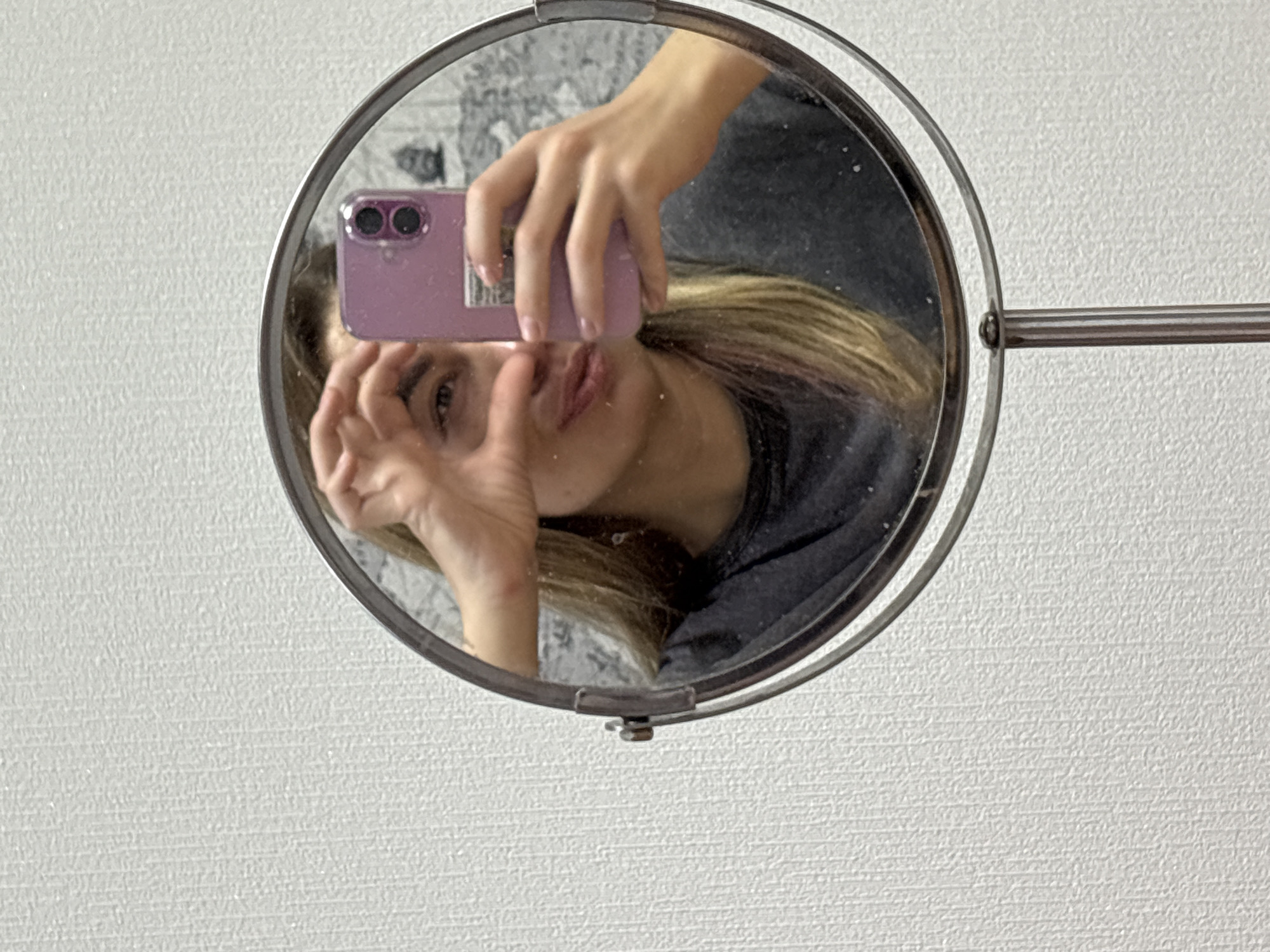 A casual mirror selfie captures an individual indoors in Ust-Ilimsk, Russia. The central element of the image is a circular mirror with a polished metal frame, reflecting the upper body of a person. The individual, with long light brown or blonde hair, is wearing a dark top. She holds a purple smartphone, featuring two prominent camera lenses, in her right hand to take the photo. Her left hand is raised near her face, with fingers slightly splayed, possibly in a playful or expressive gesture. Her lips are slightly puckered, and one eye is visible, looking directly towards the camera's reflection.
The mirror itself is mounted on a slender metallic arm that extends horizontally from its right side. The background outside the mirror is a light-colored, textured wall, likely wallpaper. Inside the mirror's reflection, the background behind the person appears darker and somewhat abstract, possibly reflecting another part of the room. The lighting suggests an indoor setting during the daytime. No specific text is clearly legible on any object in the image, although a small, indistinct sticker or label might be present on the back of the phone case. The image conveys a personal, intimate moment of self-documentation.