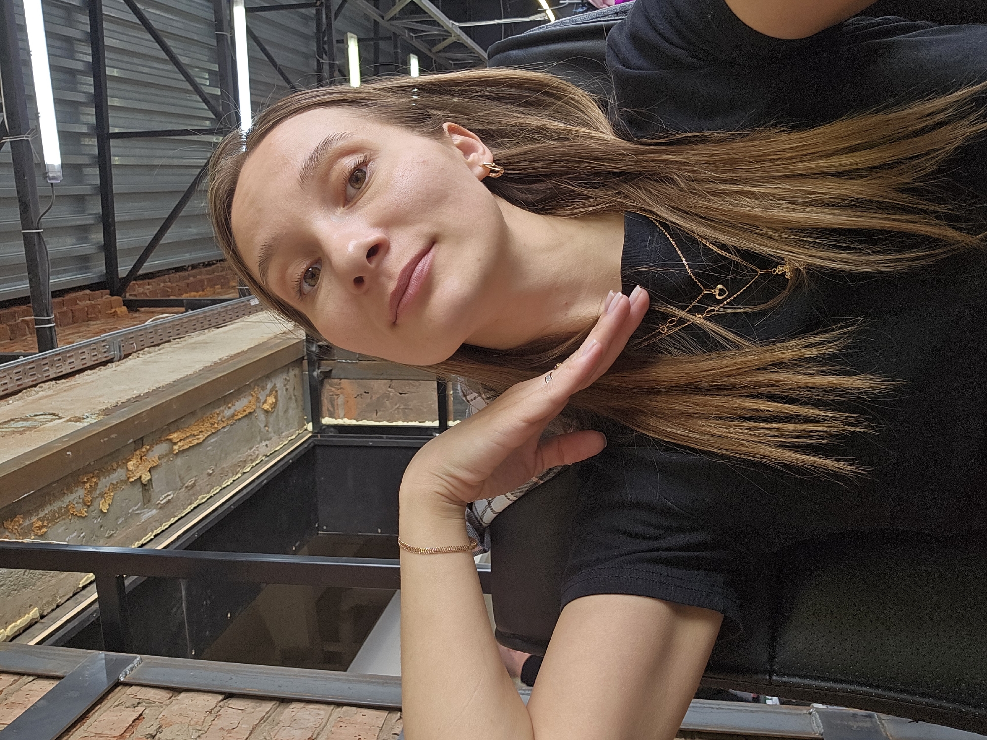 A female subject is depicted in a close-up, chest-up shot within an indoor commercial or industrial-style setting in Chaikovsky, Russia. The subject has fair skin, light brown hair extending past her shoulders, and brown eyes. She wears a plain black short-sleeved t-shirt, gold-colored hoop earrings, a thin gold-colored chain bracelet on her right wrist, and two layered gold-colored necklaces, one featuring a small heart pendant and the other a cross-like pendant. Her right hand is positioned with fingers slightly spread and angled towards her upper chest/neck area. She exhibits a subtle, closed-mouth smile, looking directly into the camera.
The background reveals a brick wall on the left side, adjoined by dark metal structural elements. Overhead, a corrugated metal ceiling is visible, supported by dark metal beams, with several rectangular fluorescent light fixtures mounted. Exposed building materials include a concrete beam exhibiting areas of dried, yellowish spray foam or decaying material. The subject appears to be seated in a black chair with a perforated texture on its backrest.