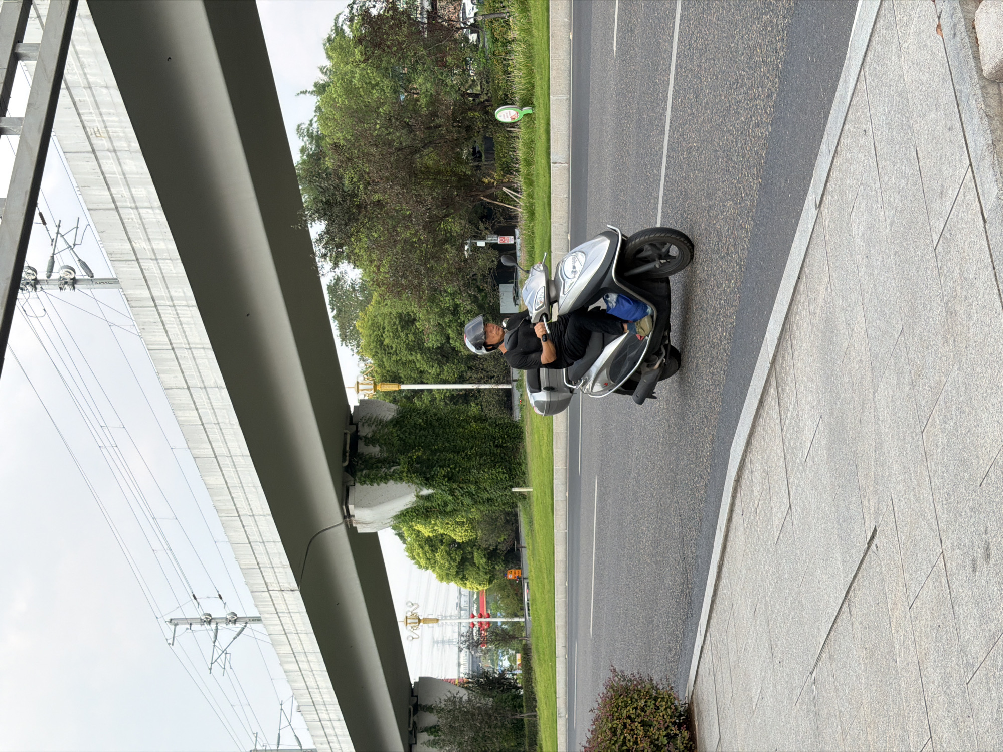 In Jiangjin, China, a man wearing a black t-shirt, dark pants, and a helmet with a clear visor rides a silver and black scooter on an asphalt road. The scooter has a black storage box mounted on the back. A blue bag is visible on the right side of the scooter. The man is looking forward, and his hands are on the handlebars. The road is bordered by a grey concrete sidewalk on the left, which is tiled with rectangular granite pavers. To the right of the road is a grassy median with some bushes and trees. In the background, a large elevated concrete structure, likely part of an overpass or elevated railway, spans across the scene. Overhead power lines and supporting poles are visible above the concrete structure. The sky is overcast. The overall environment appears to be an urban or suburban setting with infrastructure for transportation.