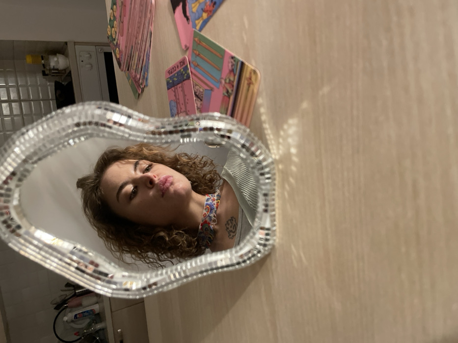 The image depicts an indoor scene in Boreti, Montenegro, featuring a wavy, mosaic-framed mirror placed on a light-colored wooden table. Reflected in the mirror is the upper body of a young adult female with curly brown hair, a patterned beaded choker, and a light-colored ribbed top. She has a slight pout and is looking downwards. A tattoo, resembling a lotus flower, is visible on her upper chest. The reflection also shows a kitchen background with white tiled walls, a counter holding various items including bottles and containers, and what appears to be a stove or oven unit. To the right of the mirror, on the table, several colorful tarot cards with dominant pink and yellow hues are spread out, some stacked and others partially fanned. The scene is illuminated by an apparent light source from the left, casting glints and reflections on the mirror frame and table surface. No direct interaction between individuals is observed.