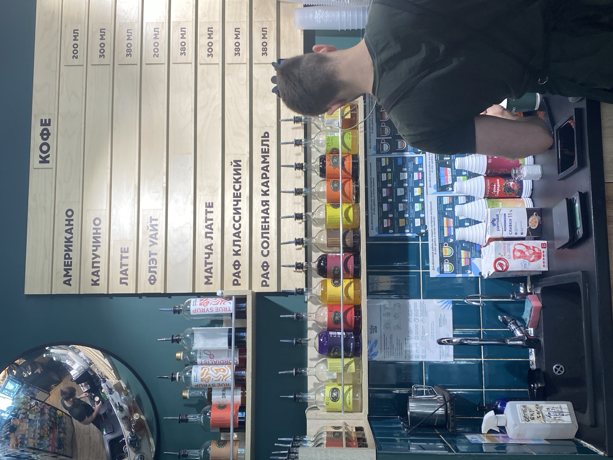 The image depicts the interior of a coffee shop or cafe, likely in Yekaterinburg, Russia, judging by the Cyrillic text on the menu. A person, viewed from behind, is standing at the counter, seemingly preparing a drink. The menu board lists various coffee types (Americano, Cappuccino, Latte, Flat White, Matcha Latte, Classic Raf, Salted Caramel Raf) along with their corresponding sizes in milliliters. Below the menu are shelves containing bottles of syrups, arranged in rows. Various ingredients and supplies, such as milk, syrups, and cleaning agents, are visible on the counter near a sink with a faucet. The wall behind the counter is decorated with teal blue tiles. A round mirror is visible on the left side of the image, reflecting the interior of the cafe, including shelves stocked with products.