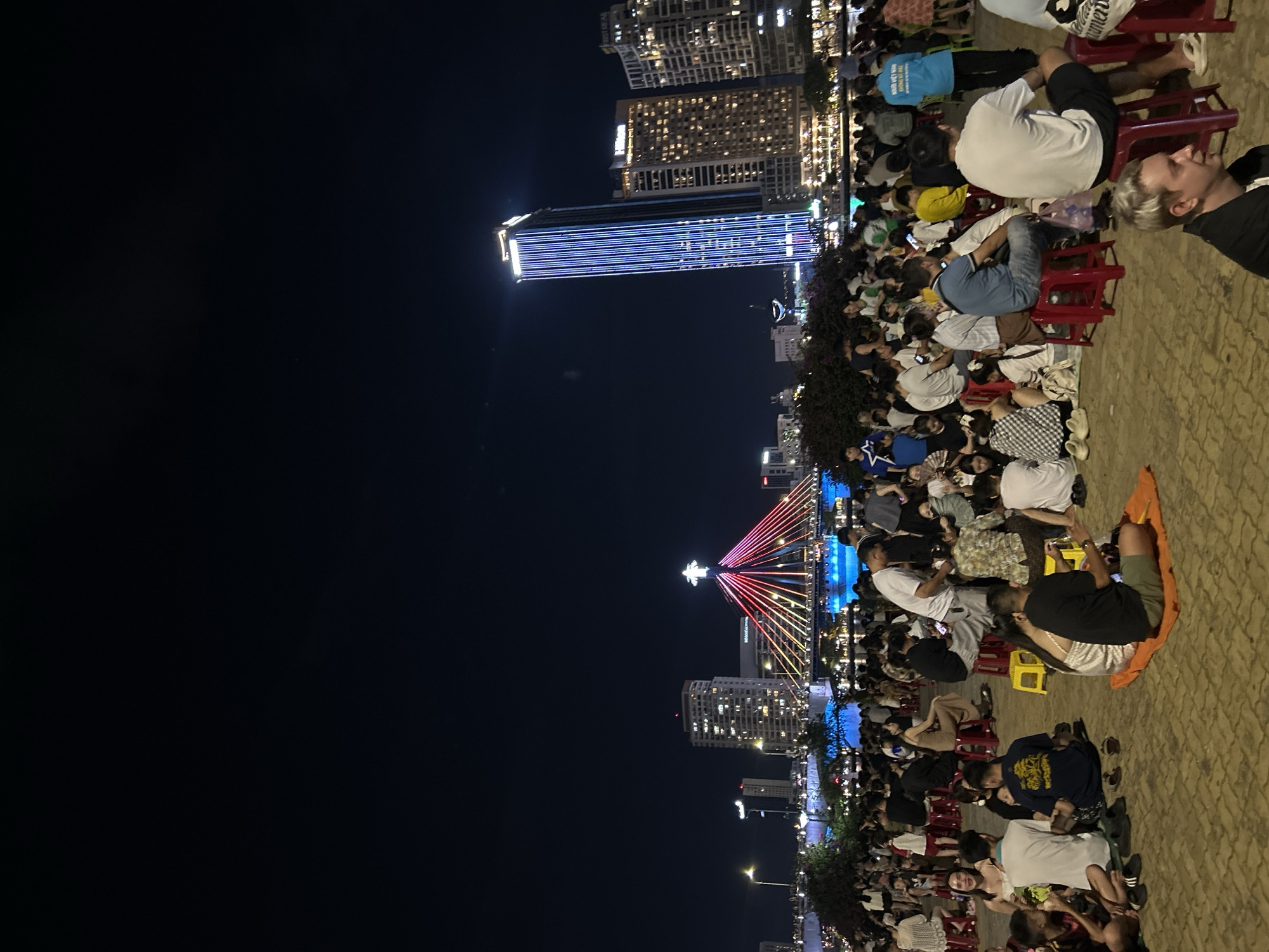 This image depicts a nighttime scene along the Han River in Đà Nẵng, Vietnam. A large gathering of people is present on a paved promenade in the foreground and midground. The individuals are predominantly adults, seated on small red or yellow plastic stools, or directly on the ground using mats or blankets. Many are engaged in conversation, observing the surroundings, or interacting with mobile phones.
In the background, across the Han River, the cityscape is brightly illuminated. The prominent Han River Bridge is visible, adorned with red, yellow, and blue lights and topped with a white star. To the right of the bridge, a tall, modern skyscraper featuring vertical blue lighting stands out, with a "Hilton" sign visible on an adjacent building to its left. Other city lights and buildings line the riverbank. Lush green foliage separates the promenade from the river. The overall atmosphere indicates a public gathering or event during evening hours.