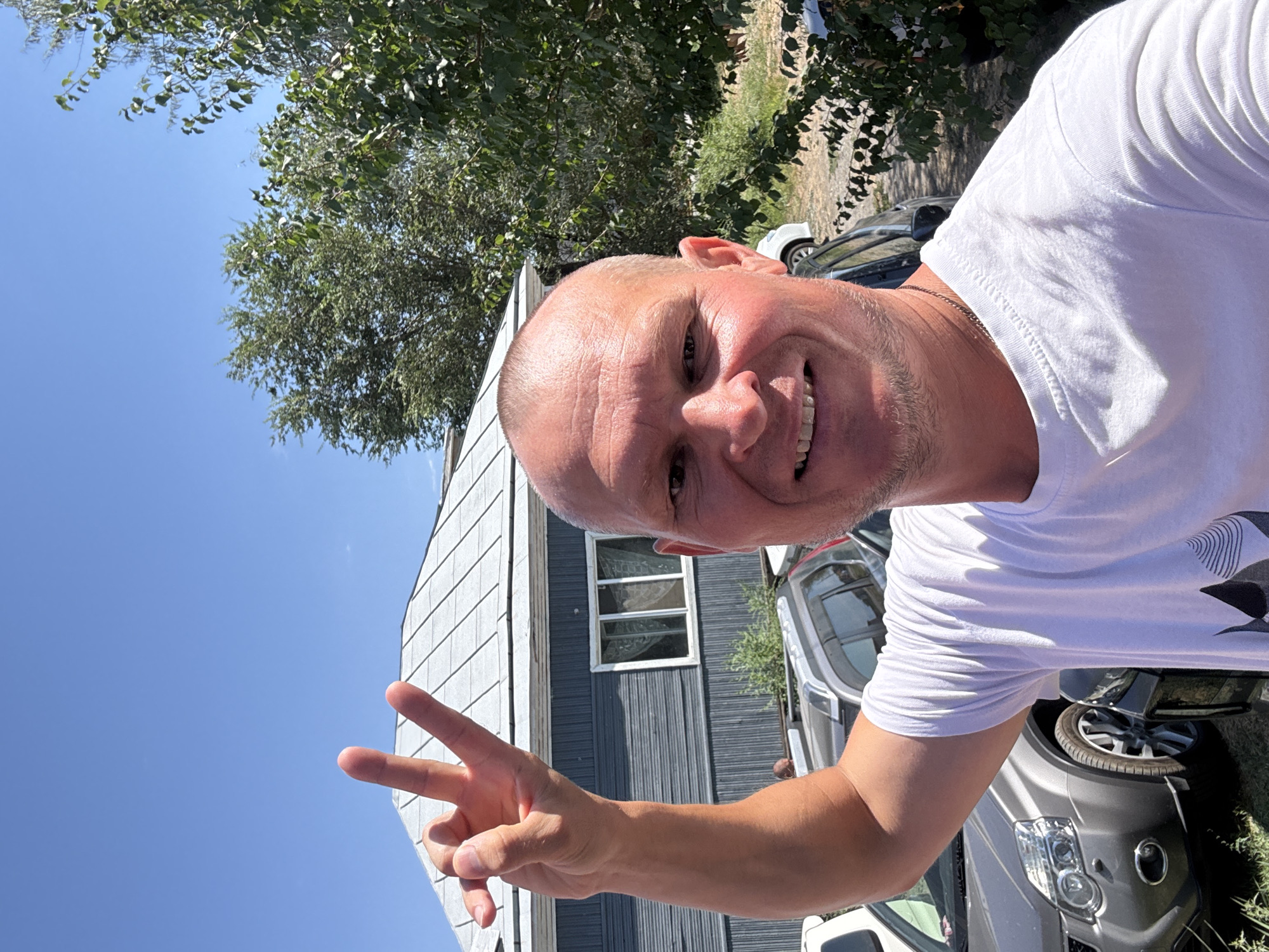 A bald, fair-skinned man in his 40s, wearing a white t-shirt, smiles at the camera, holding up his right hand with his index and middle fingers extended in a peace sign. He has a light stubble on his chin and a chain around his neck. The man is standing outdoors, with a blue sky and green trees in the background. Behind him is a gray, single-story house with a blue siding and a white-shingled roof. Several cars are parked in front of the house, including a silver SUV. The scene appears to be in a residential area.