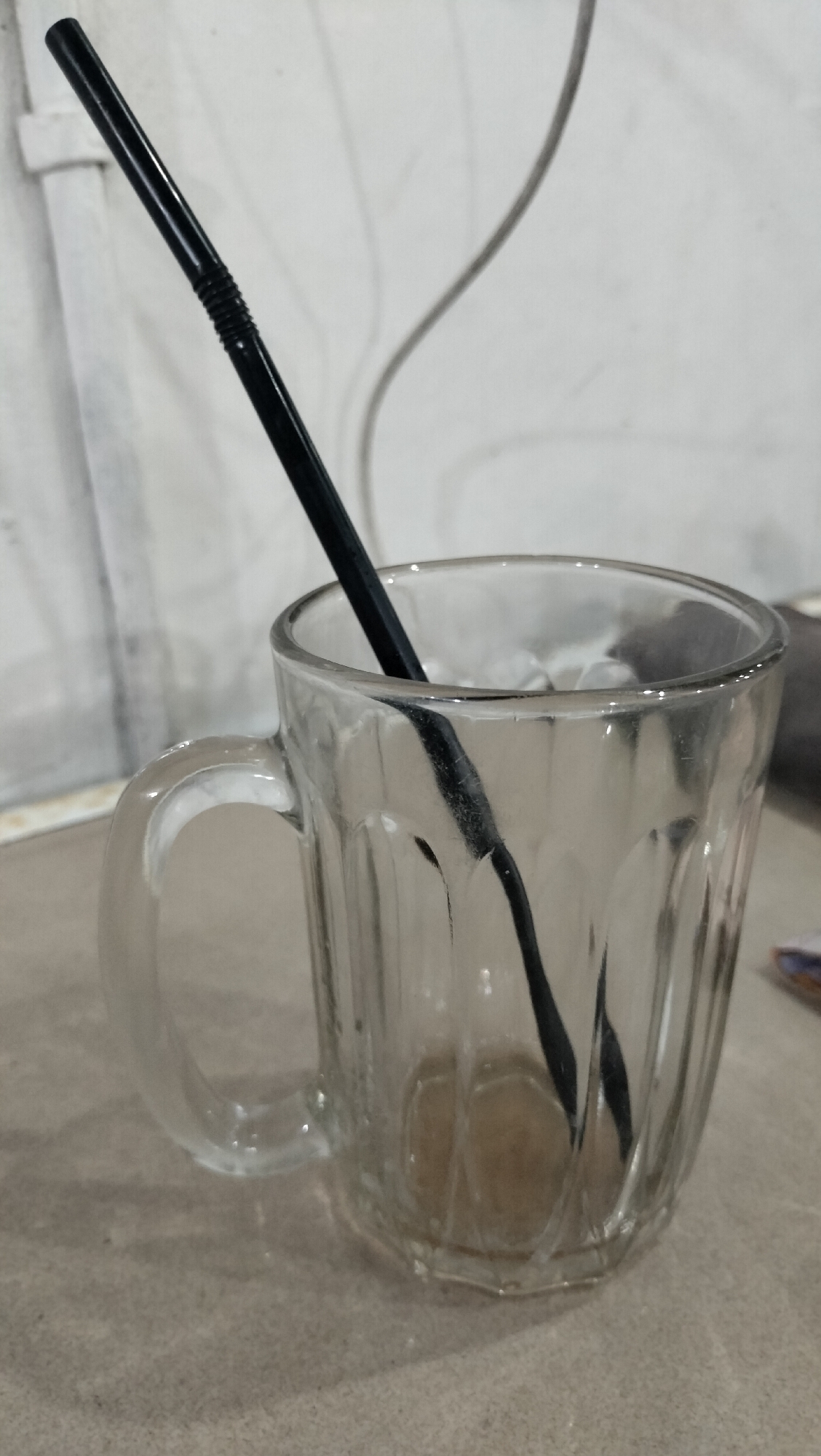 A clear, fluted glass mug with a C-shaped handle is positioned on a light gray, granular surface in Lhokseumawe, Indonesia. A black plastic straw, featuring a corrugated flexible segment, is inserted into the mug. The lower end of the straw is submerged in a small volume of light brown liquid residue at the bottom of the glass. The interior surfaces of the mug exhibit minor smudges and condensation-like patterns. The background consists of a plain, light-colored, possibly off-white, vertical surface displaying subtle linear textures. A thin, dark, curved object, consistent with a cable or wire, is visible against the background in the upper right quadrant. No human subjects are present.