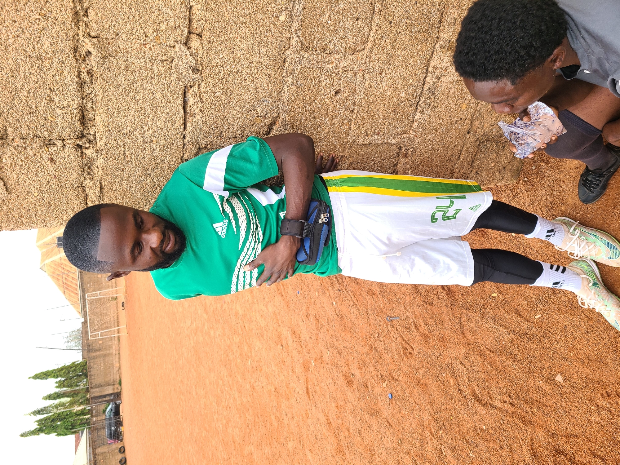 The image shows two African men on a reddish-brown, possibly dirt, field in Barakin Lerawi, Nigeria. One man is leaning against a tan brick wall, wearing a green and white Adidas jersey with the number 12 on white shorts with black knee-high socks. He has a black pouch strapped around his midsection. The second man is kneeling, hunched over an object in a foil wrapper. He is wearing a grey shirt, black shorts, and black shoes. Behind them are trees, a building, and a black car. A soccer goal net is visible in the background.