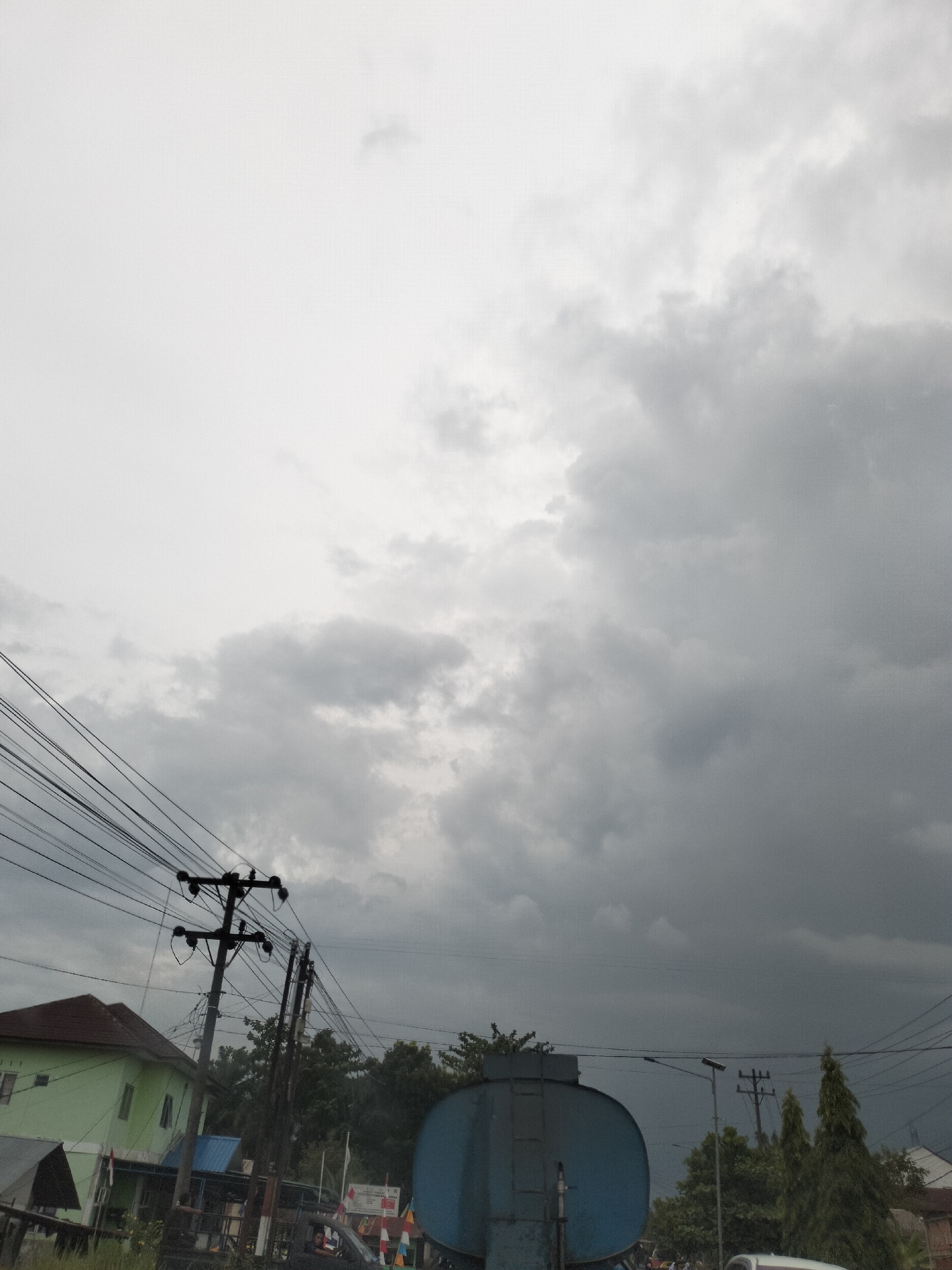 This image captures a street scene in Tangkahan Durian, Indonesia, under a heavily overcast sky. Power lines crisscross the frame, supported by wooden utility poles. To the left, a light green building with a brown roof is visible, partially obscured by a smaller gray-roofed structure. In the center, a large blue cylindrical tank truck is present. A few trees can be seen in the background, with some appearing conical and others more rounded. The overall atmosphere suggests impending or recent rain due to the dark, cloud-filled sky.