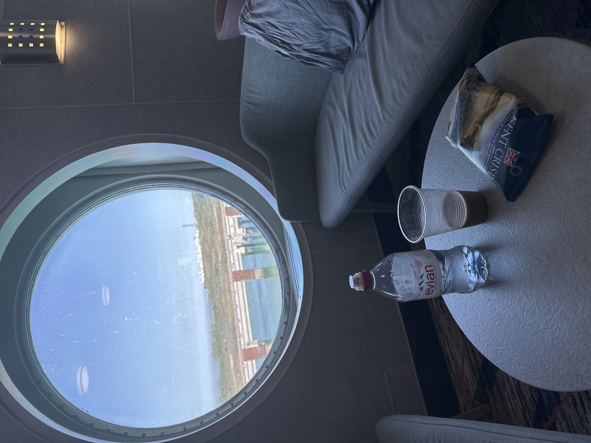 The image depicts an interior scene, likely aboard a ferry or ship, as evidenced by a circular porthole window offering a view of what appears to be the coastline of Loon-Plage, France. The view through the window shows a blue sky with some cloud cover, and land with vegetation along the shoreline. Man-made structures are also visible, including rectangular buildings and other architectural features.
Inside the space, there is a grey sofa with a crumpled grey fabric on it. Next to the sofa, there is a small round table. On the table, there is a bottle of "Evian" water, an empty drinking glass, and a package of "Kent Crisps" sea salt and vinegar potato chips. A light fixture is visible on the wall.