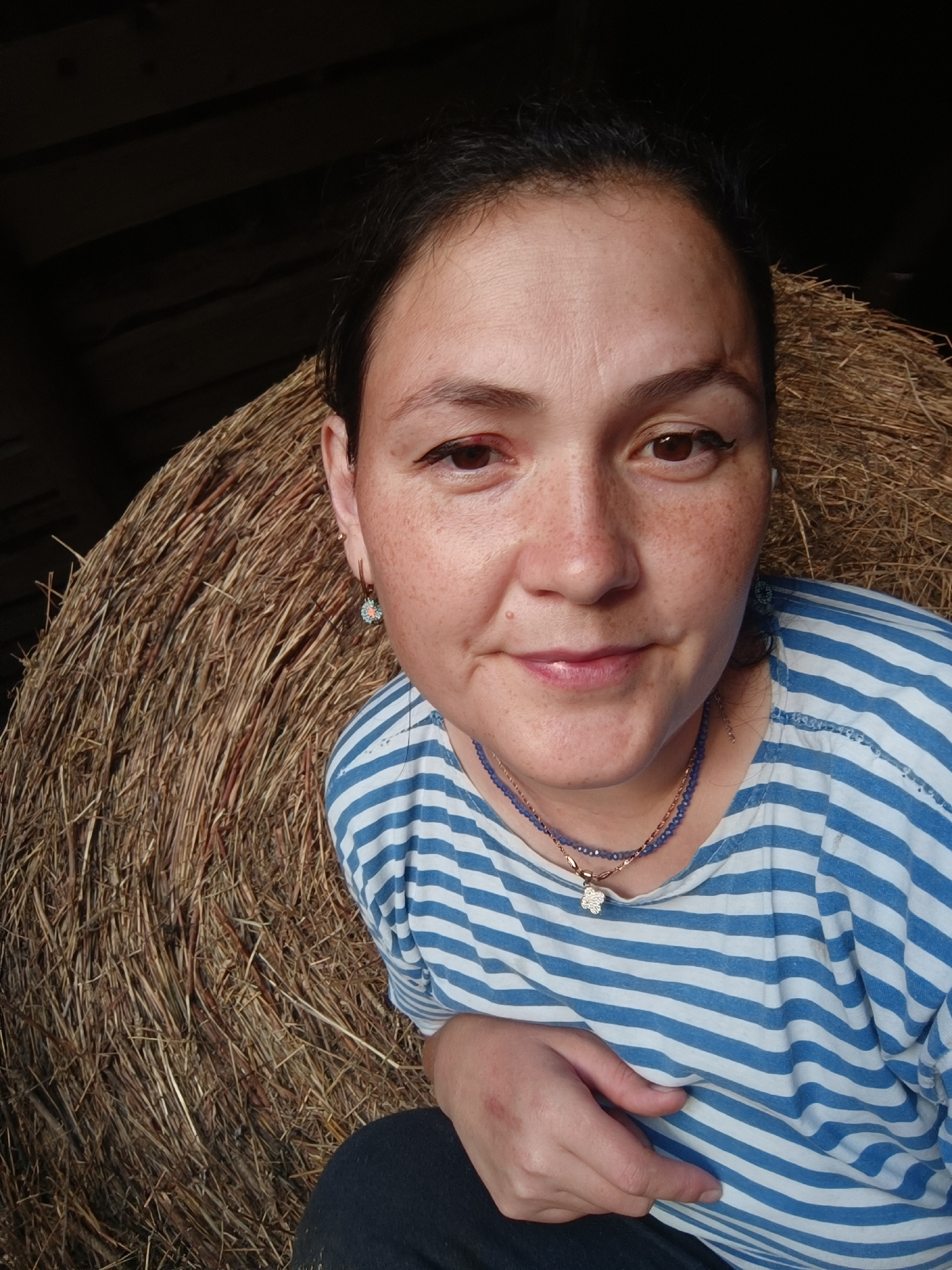 A woman is pictured in a close-up selfie, seated in front of a large haystack. She has brown hair pulled back and freckles across her face. Her right eye appears slightly swollen. She is wearing a blue and white striped t-shirt and a blue beaded necklace with a flower pendant. Her left hand is resting on her chest. The background suggests a rustic setting, possibly a barn, with dark wooden beams visible. The lighting is warm, with a shadow cast from the left side of the frame.