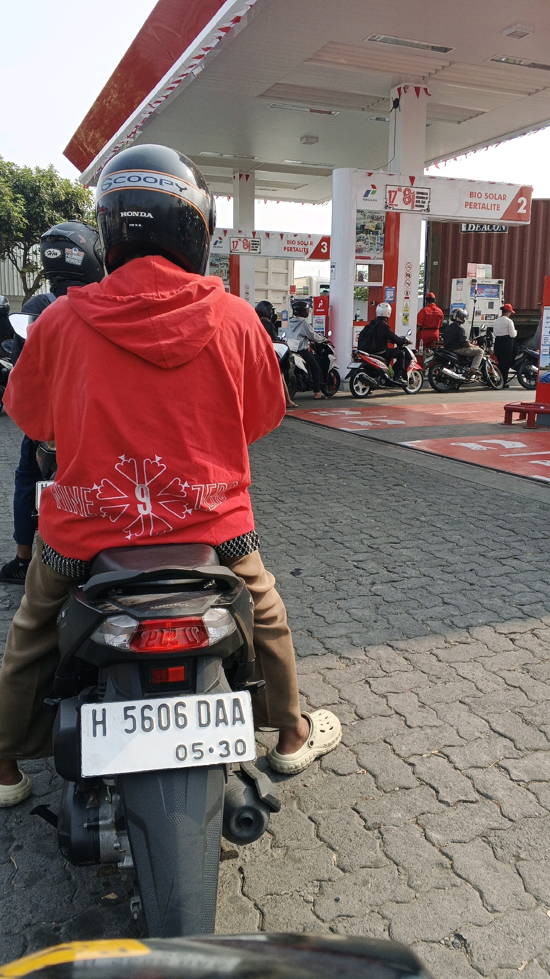 A rear view of a person on a motorcycle at a gas station in Semarang, Indonesia. The person is wearing a red hooded jacket with a white graphic design and light brown pants, and cream-colored clogs. The motorcycle has a white license plate that reads "H 5606 DAA 05.30". In the background, there are several other motorcycles and people at the gas pumps under a red and white striped canopy. The ground is paved with grey interlocking bricks.
