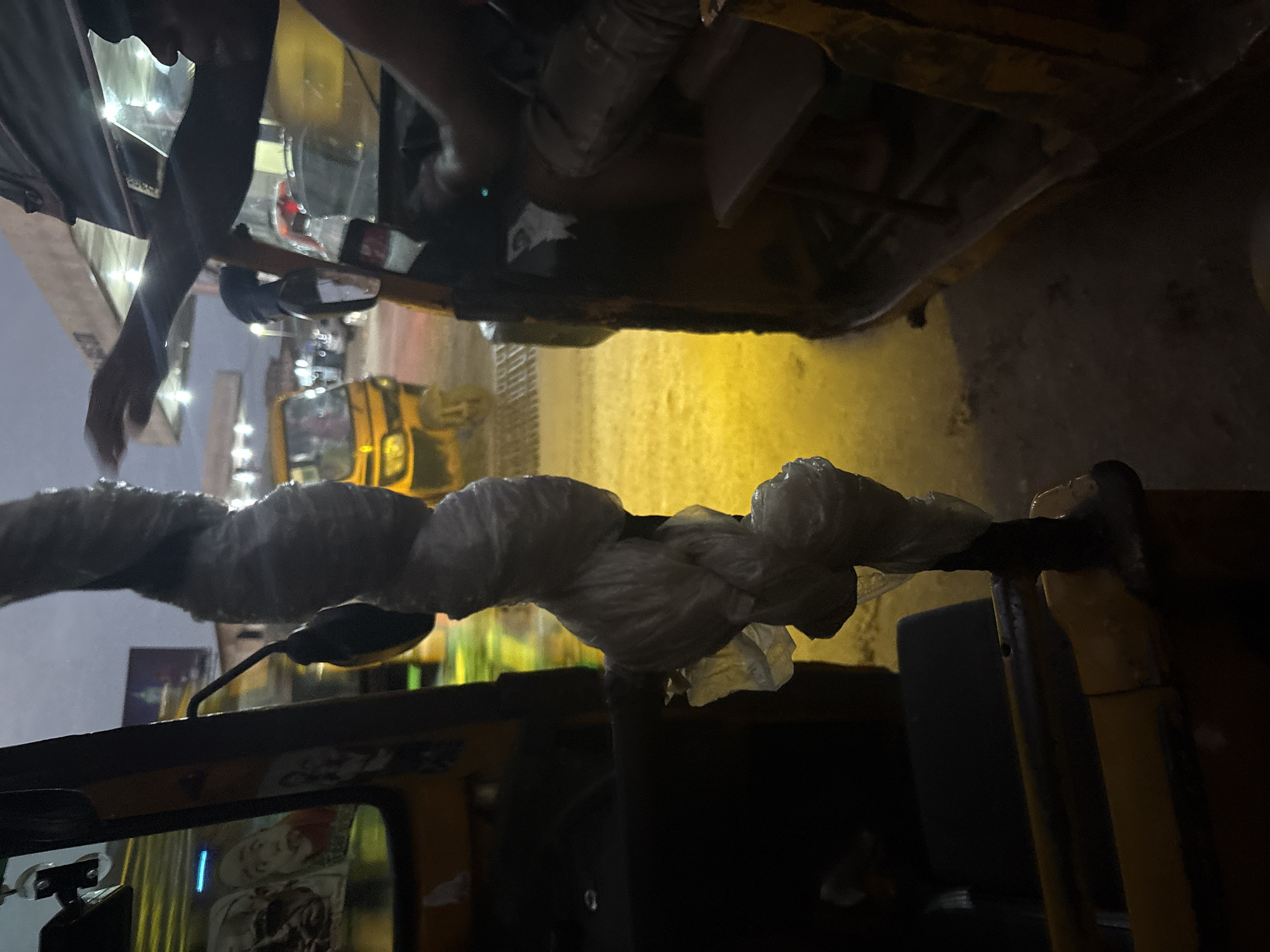 This image captures a scene on a street in Onitsha, Nigeria, likely at dusk or night, indicated by the dim lighting and artificial illumination. The foreground is dominated by the interior of a yellow auto-rickshaw, with a thick, white-wrapped cable or pipe obscuring the view. To the right, another auto-rickshaw is partially visible, with its interior in darkness, except for what appears to be a passenger seat and the arm of a person.
In the background, another yellow auto-rickshaw is more clearly visible, its headlights on, parked on a wet, illuminated street. The yellow light reflects off the wet surface, suggesting recent rain. The overall environment appears to be a busy urban setting, typical of a commercial hub like Onitsha, with hints of buildings and other vehicles further in the distance.
The imagery inside the first auto-rickshaw suggests a personal touch or decoration, with what look like printed images or stickers on the window. The atmosphere is somewhat hazy, possibly due to humidity or the quality of light. The focus is on the immediate surroundings within the vehicle and the street scene unfolding just beyond it.