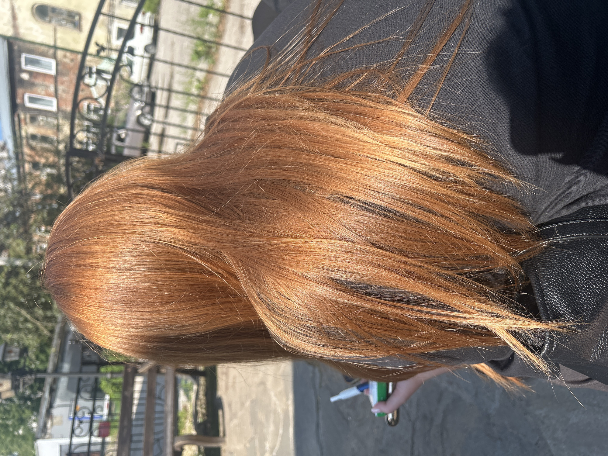 The image shows a person from behind, with their long, reddish-brown hair prominently displayed. The person is wearing a black jacket and holding a small object in their left hand that appears to be a vape pen with a white tip and a green casing. The background suggests an urban outdoor setting, possibly a park or a public square. There are hints of a bench, a fence, a building with multiple windows, and cars in the distance. The lighting indicates it is daytime, with shadows suggesting sunlight. Based on the style of the buildings and the overall atmosphere, the scene could be located in Perm, Russia, particularly an older part of the city.