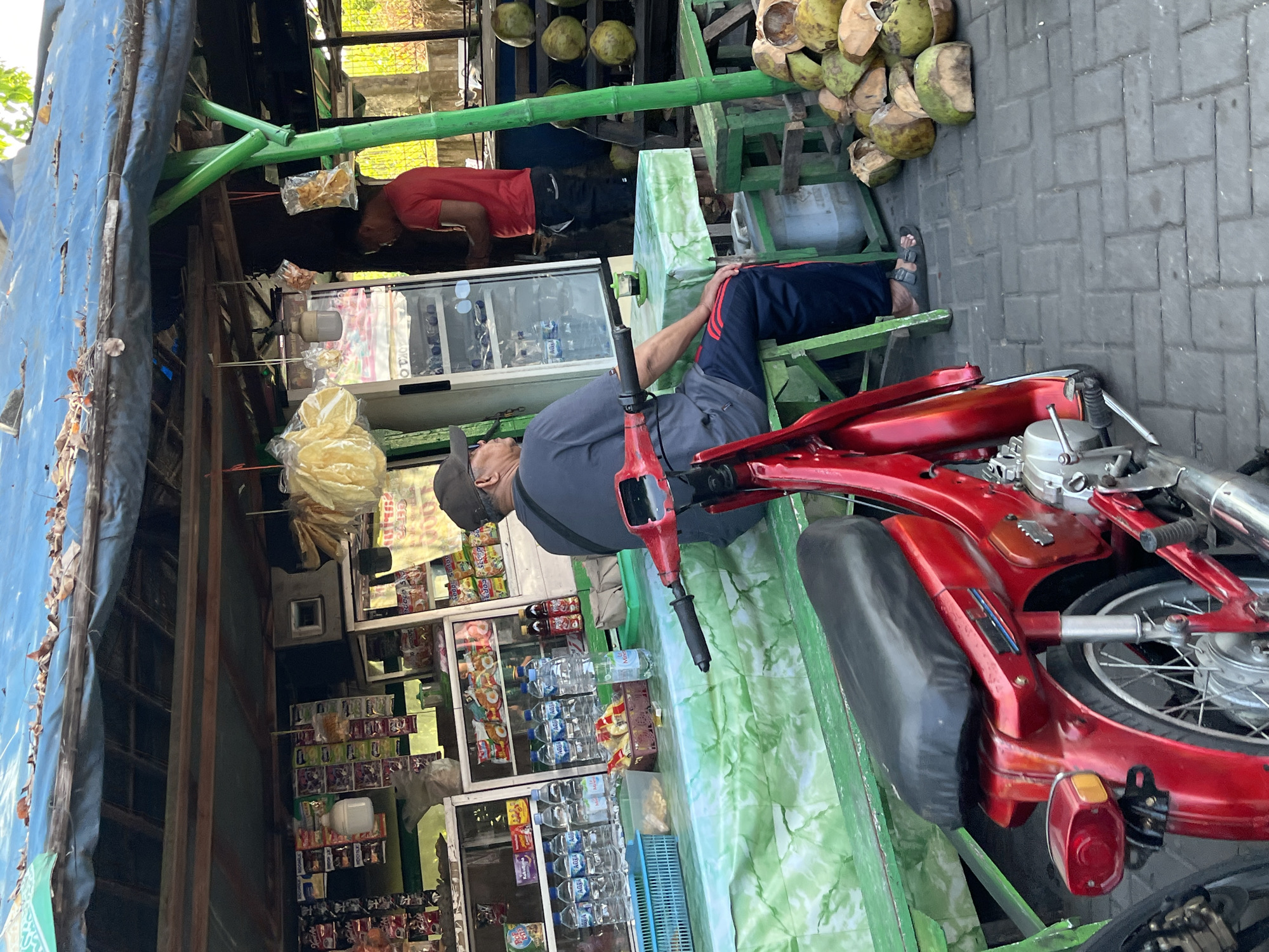 The image depicts a street-side refreshment stall in Juwet Kenongo, Indonesia. A man wearing a dark blue t-shirt and dark pants with red stripes sits on a green bench in front of the stall. He is facing away from the camera, looking towards a small table with a cup of dark liquid. Beside him, a red motorcycle is parked. Behind the man is a stall with various items for sale displayed on shelves, including bottled water, snacks, and other packaged goods. A young man in a red t-shirt stands inside the stall, near a refrigerator unit. A stack of halved coconuts is visible on the right side of the image, near a wooden stand. The stall is covered by a blue tarp. The ground is paved with grey interlocking bricks.