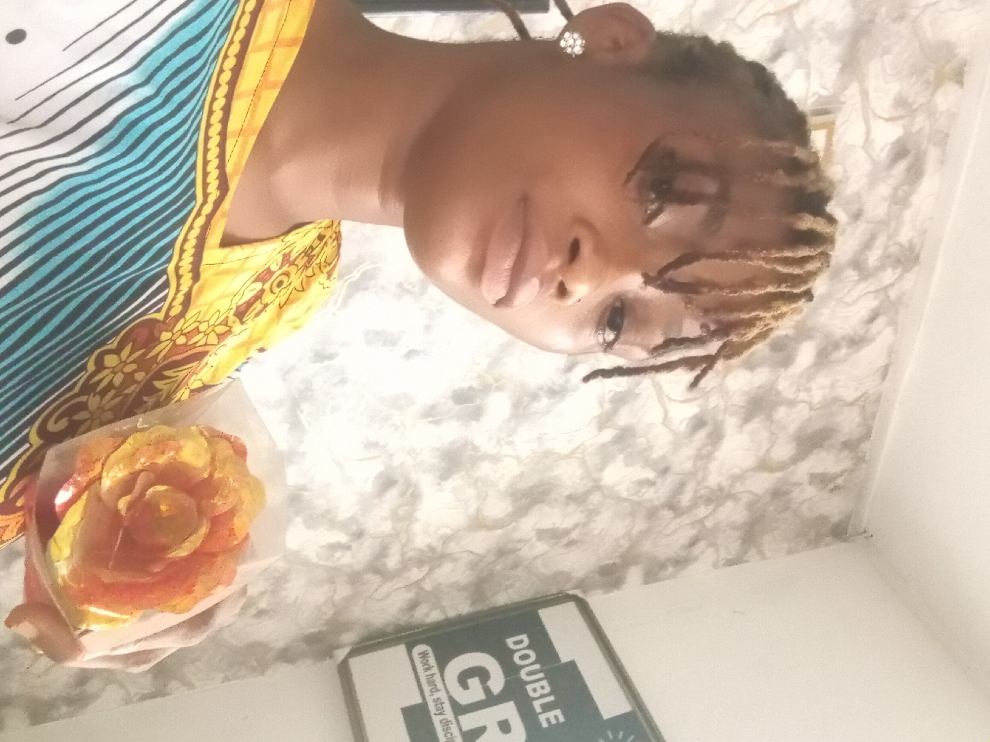 A medium-shot, high-angle photograph depicts a woman indoors holding a wrapped flower. The woman has light brown skin and wears a printed top with blue, yellow, brown and white stripes. Her hair is styled into blonde and brown braids and she wears an earring on her right ear. She looks towards the camera. She holds a rose, packaged in clear wrapping.
A white and grey patterned background appears behind the woman. In the lower portion of the photo, a green and white sign with the words "DOUBLE GR" and "Work hard, stay disciplined" is visible.
No definitive information confirms that the location of the photograph is Shagamu, Nigeria.