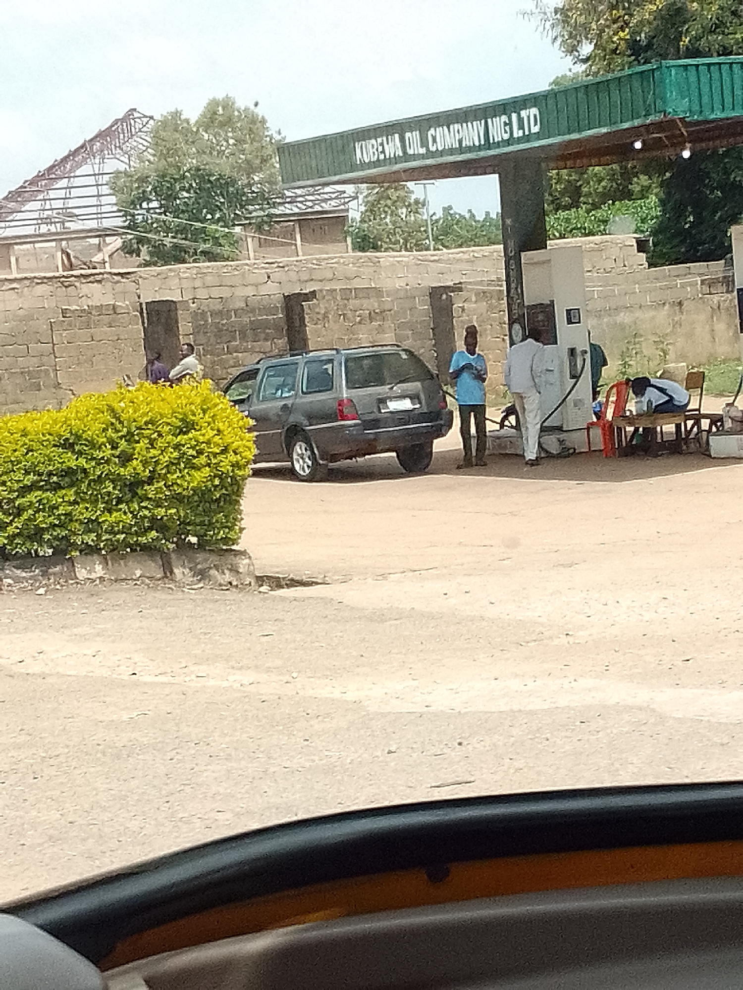 The image depicts a scene at the Kubewa Oil Company Nig Ltd gas station in Jos, Nigeria. A dark-colored hatchback car is parked at a gas pump. Several individuals are present: one man is standing next to the car in a blue shirt, and another man in a white outfit appears to be interacting with the gas pump. Another person is visible, appearing to be either sleeping or resting on a table and chair. A dense, green bush grows along the edge of the road in the foreground. A low brick wall runs along the rear of the station, behind which an incomplete building structure is visible. The foreground of the image is out of focus, suggesting the photo was taken from inside a vehicle. The environment appears dusty and dry.