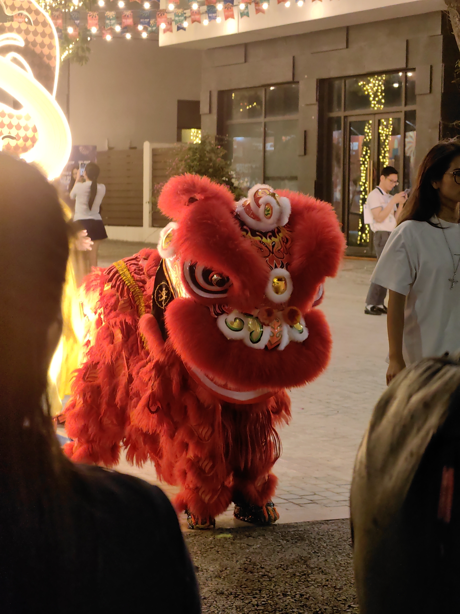 The image shows a traditional red lion dance costume in Bai Chay Ward, Vietnam. The lion's head is elaborate and brightly colored, with white, gold, and black details. The body is covered in red fur-like material. People are standing around watching and photographing the lion dance. The background includes buildings with lit windows and festive lights strung overhead. The ground is paved.