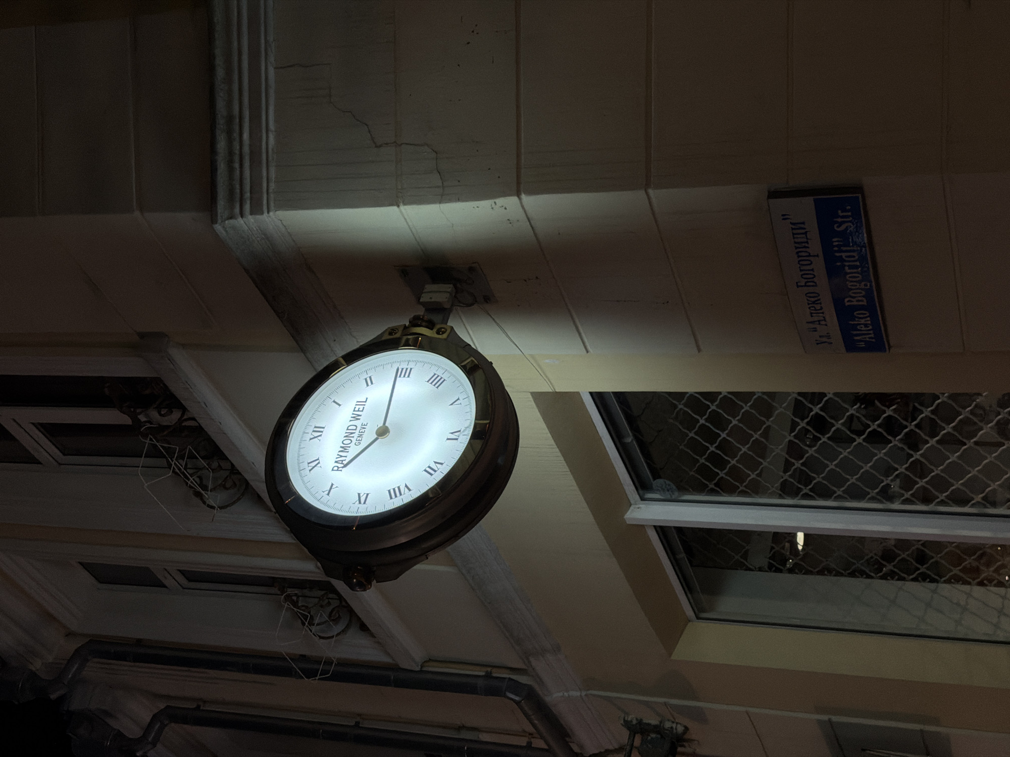 The image captures a street scene at night in Burgas, Bulgaria, focusing on a large, illuminated clock mounted on the side of a building. The clock face features Roman numerals and the "RAYMOND WEIL GENEVE" brand. Below the clock, a street sign is visible, reading "ул. „Алеко Богориди”" in Cyrillic and ""Aleko Bogoridi" Str." in English, indicating the location as Aleko Bogoridi Street. The building has a pale yellow facade with architectural details and windows. The lighting suggests it is nighttime, with the clock being a prominent light source. The environment appears to be an urban setting with no people or visible activity.