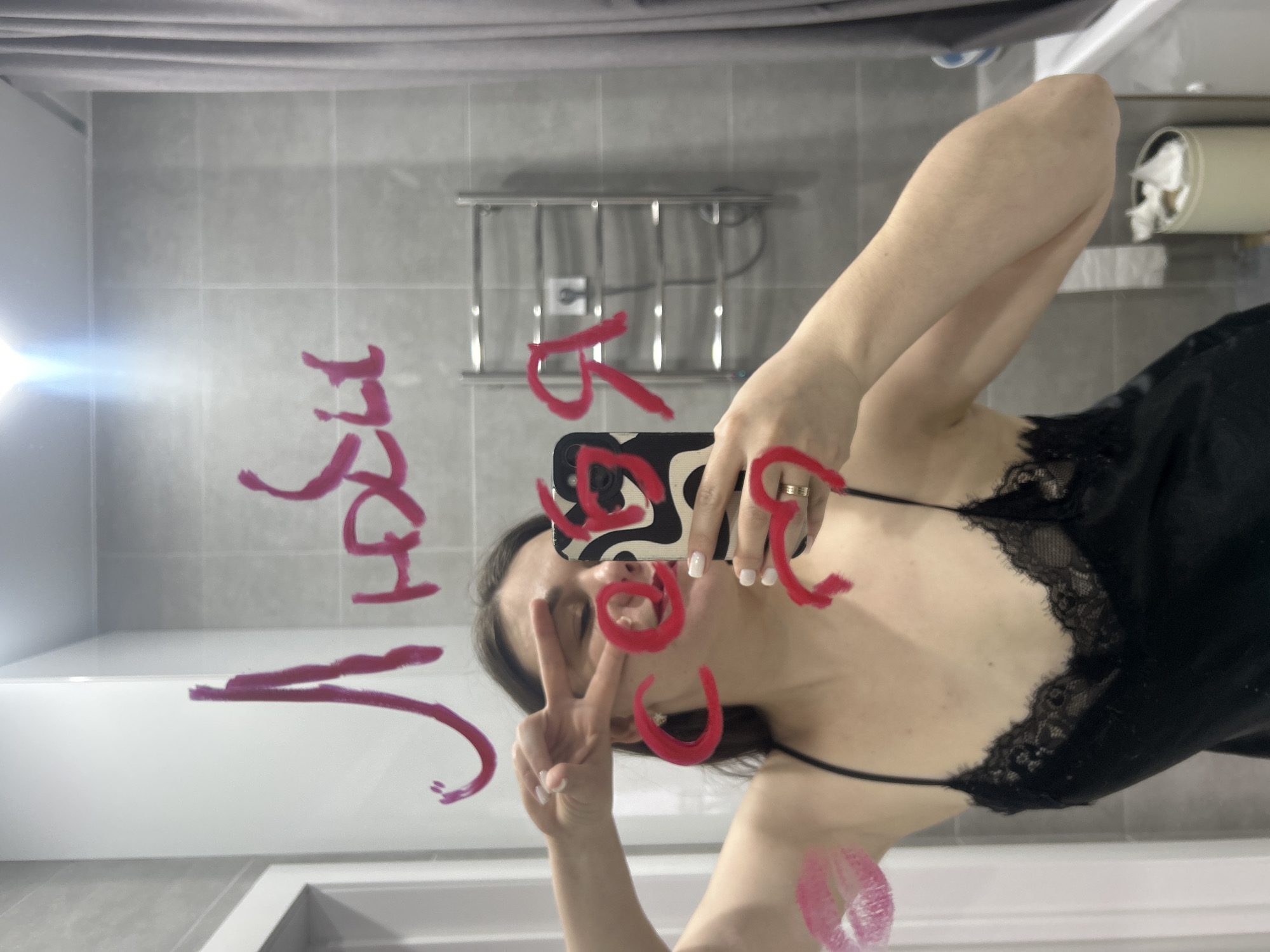A mirror selfie taken by a woman in a modern bathroom in Chaikovsky, Russia, shows her from the chest up. She has dark hair and wears a black, lace-trimmed camisole. Her right hand holds a smartphone with a black and white abstract pattern, partially obscuring her face, while her left hand forms a peace sign. She wears a ring on her left ring finger, and her fingernails are painted white.
The mirror's surface is covered with messages and symbols drawn in red lipstick. Visible in reflection, the phrase "YOU ARE HOT" is scrawled across the mirror in a casual, cursive-like style. A clear lipstick kiss mark is also present on the lower right side of the mirror.
The bathroom features light grey tiled walls and a white bathtub or sink rim in the foreground. In the background, a silver-toned towel rack or heater is mounted on the tiled wall. To the far right, a wall-mounted toilet paper holder with crumpled paper is visible. The lighting suggests an indoor setting, and the overall impression is playful and self-assured.