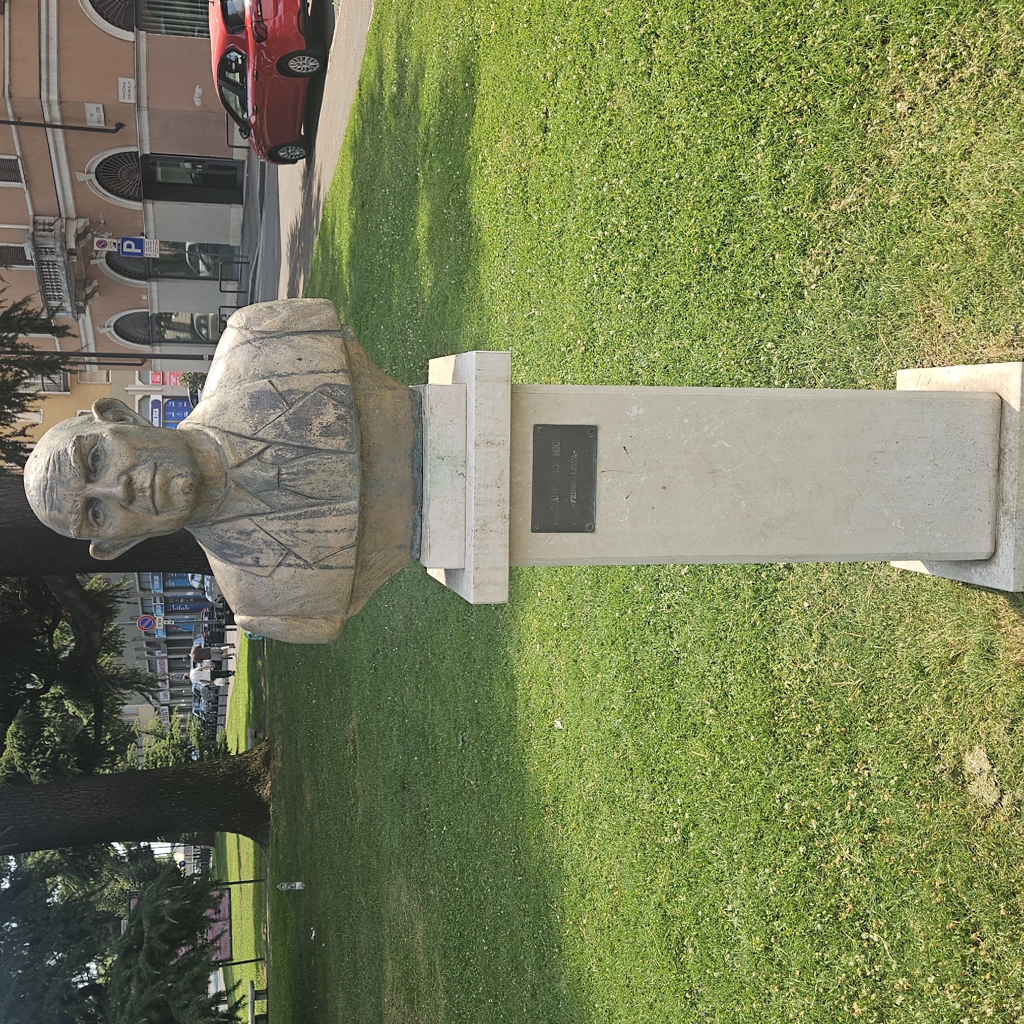 A bust of a man is displayed on a stone pedestal in a grassy park. The bust is made of a textured, light brown material and depicts a man with a bald head, prominent ears, and a stern expression. He is wearing a jacket with a patterned design on the lapels and shoulders. A dark metal plaque is affixed to the front of the pedestal. Visible text on the plaque reads: "FRANCESCO MIGLIORATO" and below it "-FEDE LIBERTA-". The park setting includes green grass, a large tree with dark bark and dense foliage to the left of the bust, and a paved walkway. In the background, across the walkway, is a building with arched windows and a red car parked in front. Further in the background, there are more buildings, street signs, and people walking. The scene appears to be set during the daytime, under sunny weather, given the shadows cast by the tree and the overall brightness of the image. The location is Brescia, Italy.