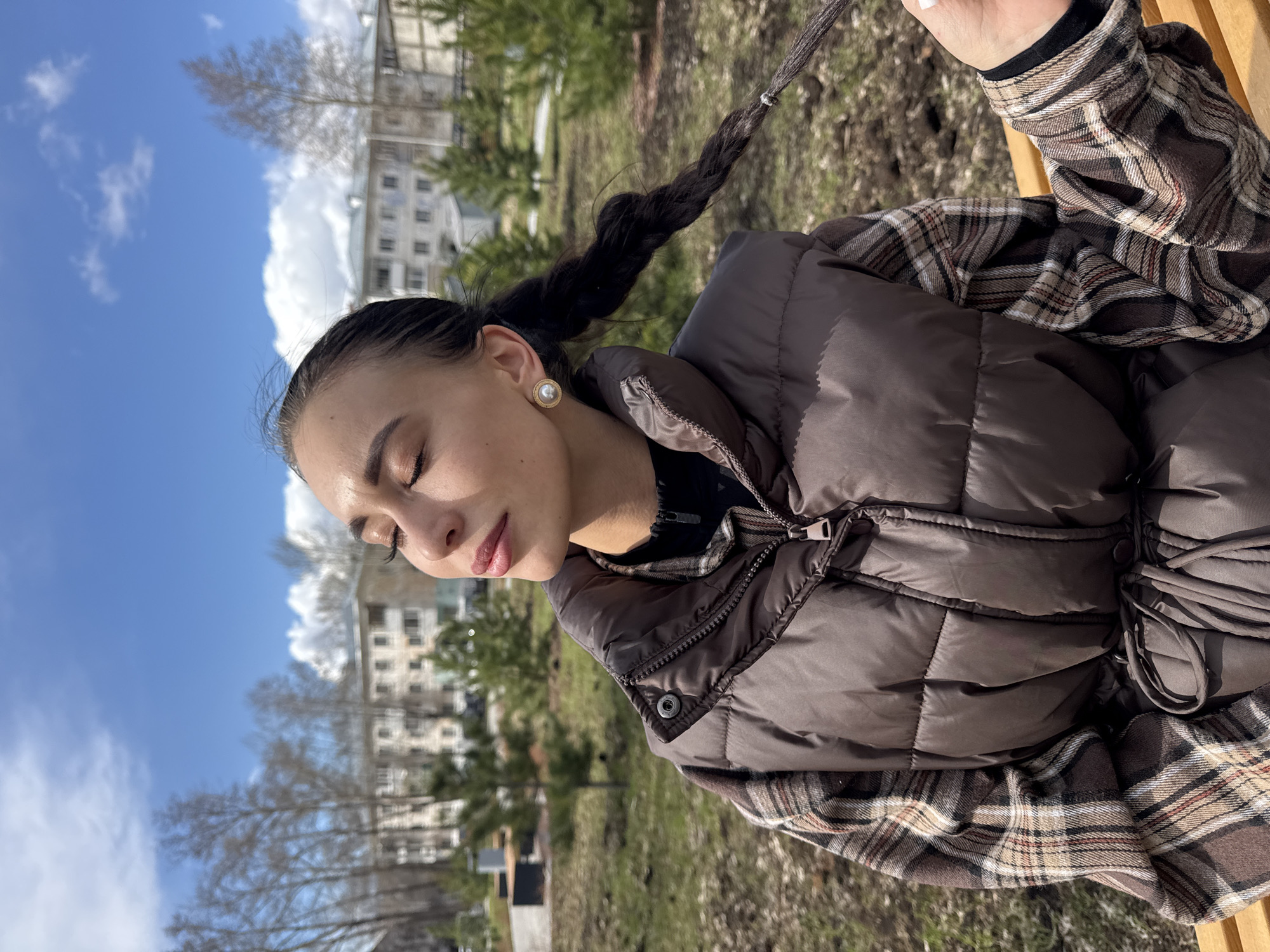 A young woman is the central subject of this image, captured outdoors in what appears to be a park or urban green space. She is turned slightly to her right, with her eyes closed and lips softly parted, conveying a sense of peace and relaxation, possibly basking in the sunlight. Her dark hair is styled into a long, thick braid that drapes over her right shoulder. She wears a quilted brown puffer vest over a plaid shirt featuring shades of brown, beige, black, and white. A prominent round pearl earring with a gold-colored rim is visible on her right ear.
The background reveals a clear blue sky dotted with scattered white clouds, suggesting pleasant weather. Below the horizon, there are trees, some appearing bare as if in late winter or early spring, and others with green foliage. In the mid-ground, several multi-story buildings are visible, indicating an urban or semi-urban environment. The ground beneath the trees is covered with grass and earth. The overall lighting suggests it is daytime, with bright sunlight illuminating the scene. No visible text is present in the image.
