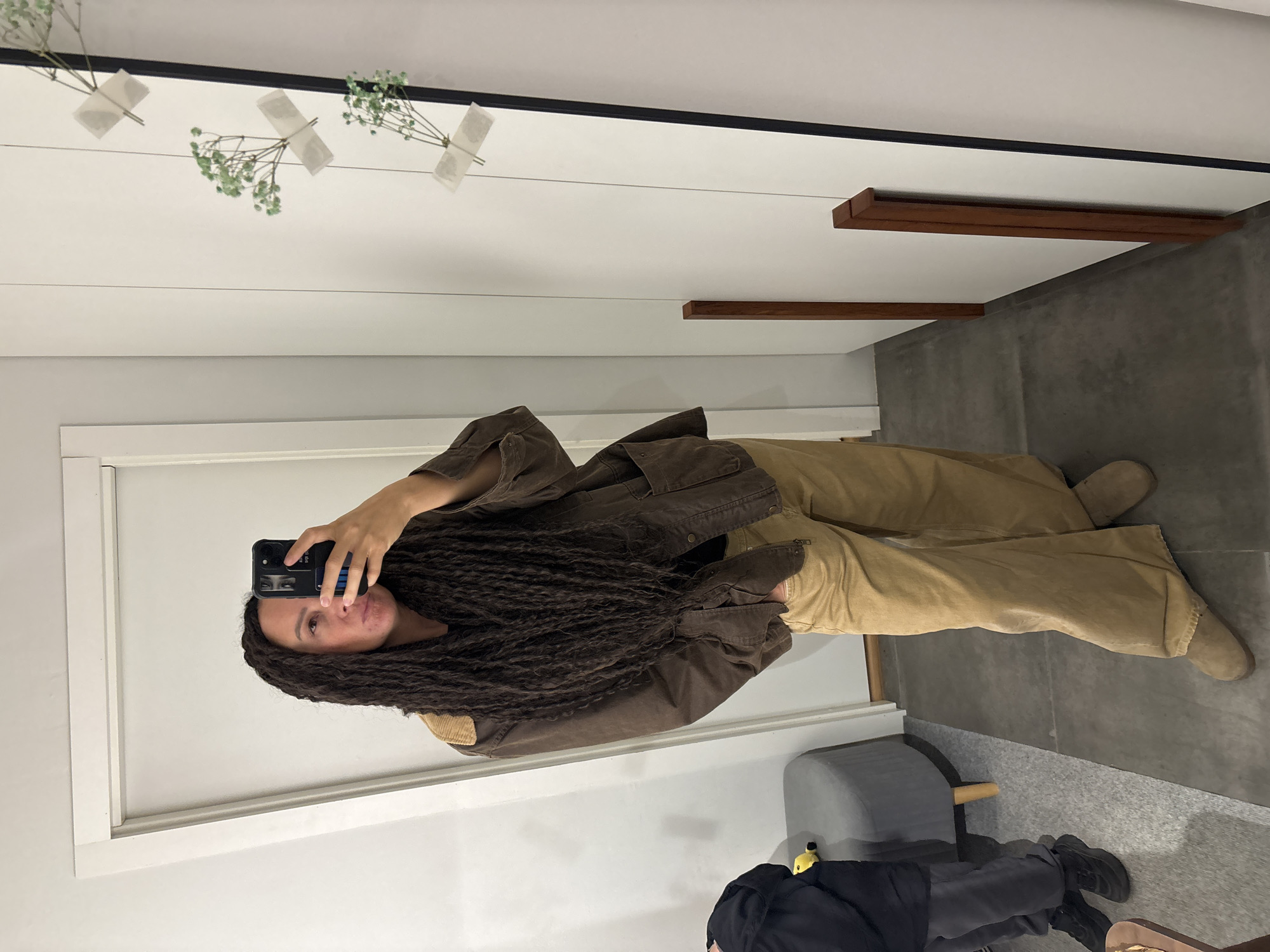 This media file captures an indoor mirror selfie, rotated 90 degrees clockwise. The main subject, a person with long, dark dreadlocks or braids, is seen taking the photograph with a smartphone. They are wearing a brown, possibly canvas or utility-style jacket with corduroy elbow patches, and loose-fitting khaki or tan pants, paired with light-colored boots. Their expression is neutral, looking towards the phone.
In the bottom right corner, partially visible, are the legs and feet of another person, clad in dark clothing and black shoes, next to a gray upholstered stool with light wooden legs. A small yellow object lies on the floor near the stool.
The setting is an interior space, characterized by a white wall with a thin black trim at the top and a white-framed mirror. Decorative elements include several small sprigs of light green foliage, possibly baby's breath, individually taped to the white wall with white tape. Two dark brown wooden bars or shelves are also mounted horizontally on the wall. The floor exhibits two distinct textures and shades of gray. On the back of the smartphone case, there is a blue vertical stripe with illegible white text. The scene is well-lit, typical of an indoor environment, with no discernible time of day.