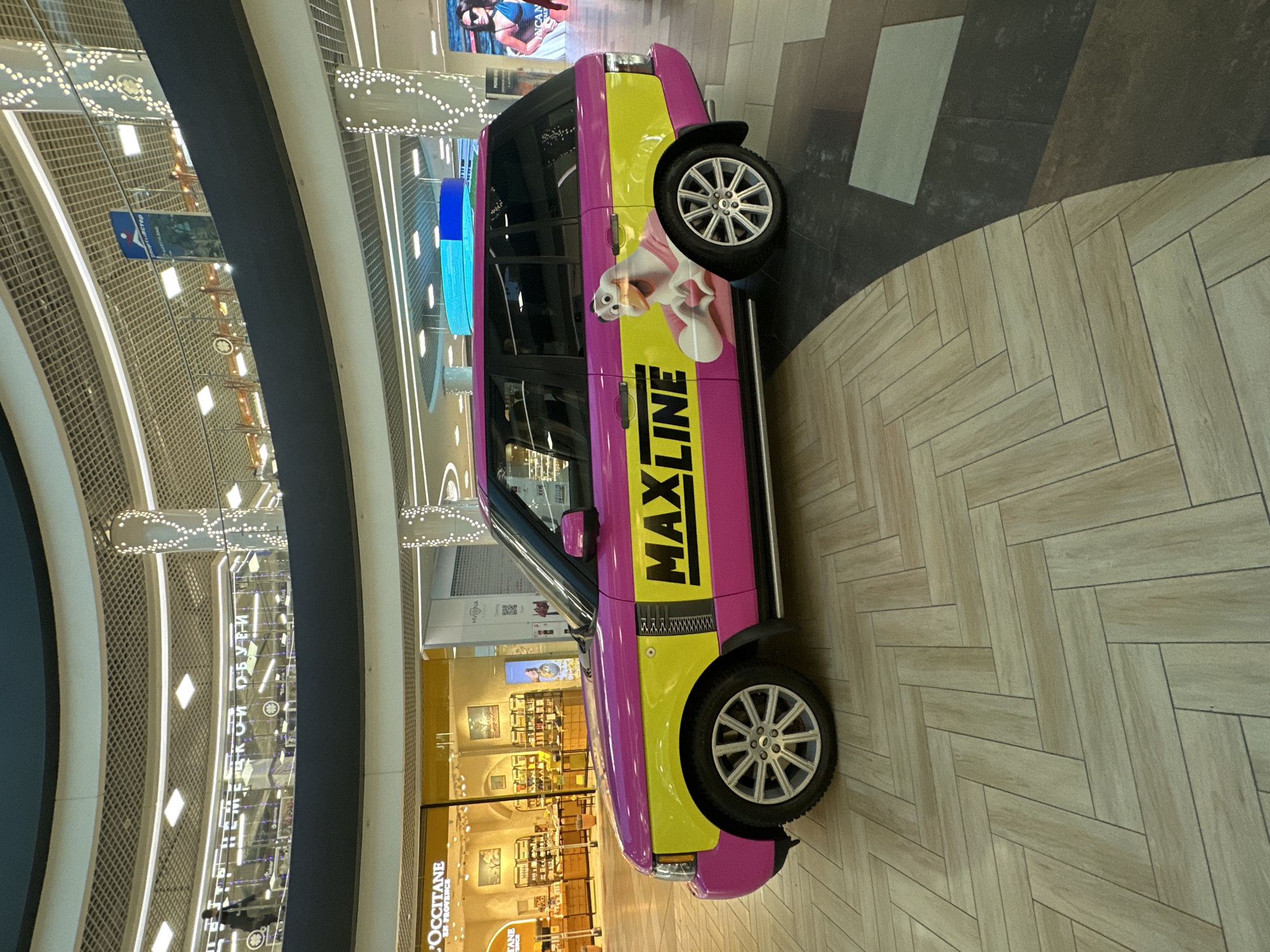 The image shows the side profile of a pink and yellow Range Rover parked on a herringbone patterned wooden floor in a modern indoor shopping mall. The vehicle is branded with "MAXLINE" in large black letters and features a cartoon duck illustration on the rear door. In the background, blurred shops are visible, including one labeled "L'OCCITANE EN PROVENCE". Circular balconies with decorative lighting are overhead, suggesting a multi-level interior. The overall environment is brightly lit, indicative of a commercial retail space. The scene is devoid of any discernible interaction between people and the vehicle or other elements.