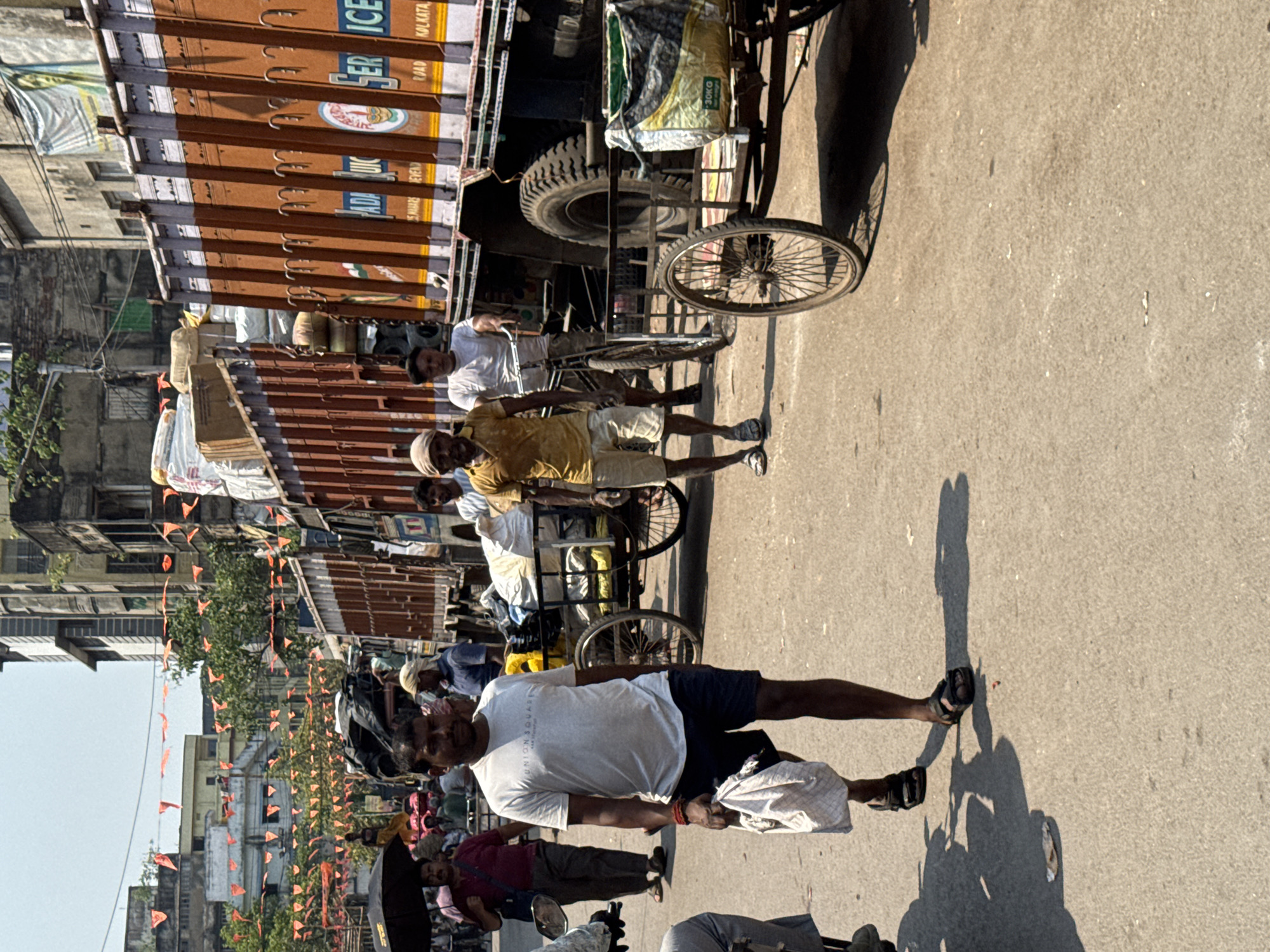 The image shows a street scene in Kolkata, India. A large truck with brown metal sides and a black top is parked on the street, with the words "SER ICE KOLKATA" visible on its side. A man in a white shirt and dark shorts is walking towards the foreground, holding a white cloth bag in his hand. Several other people are walking in the same direction. A bicycle rickshaw is positioned near the truck, with a bag on its seat. In the background, there are buildings decorated with small orange flags, suggesting a festive occasion or procession. The street is paved, and the lighting indicates it is daytime.