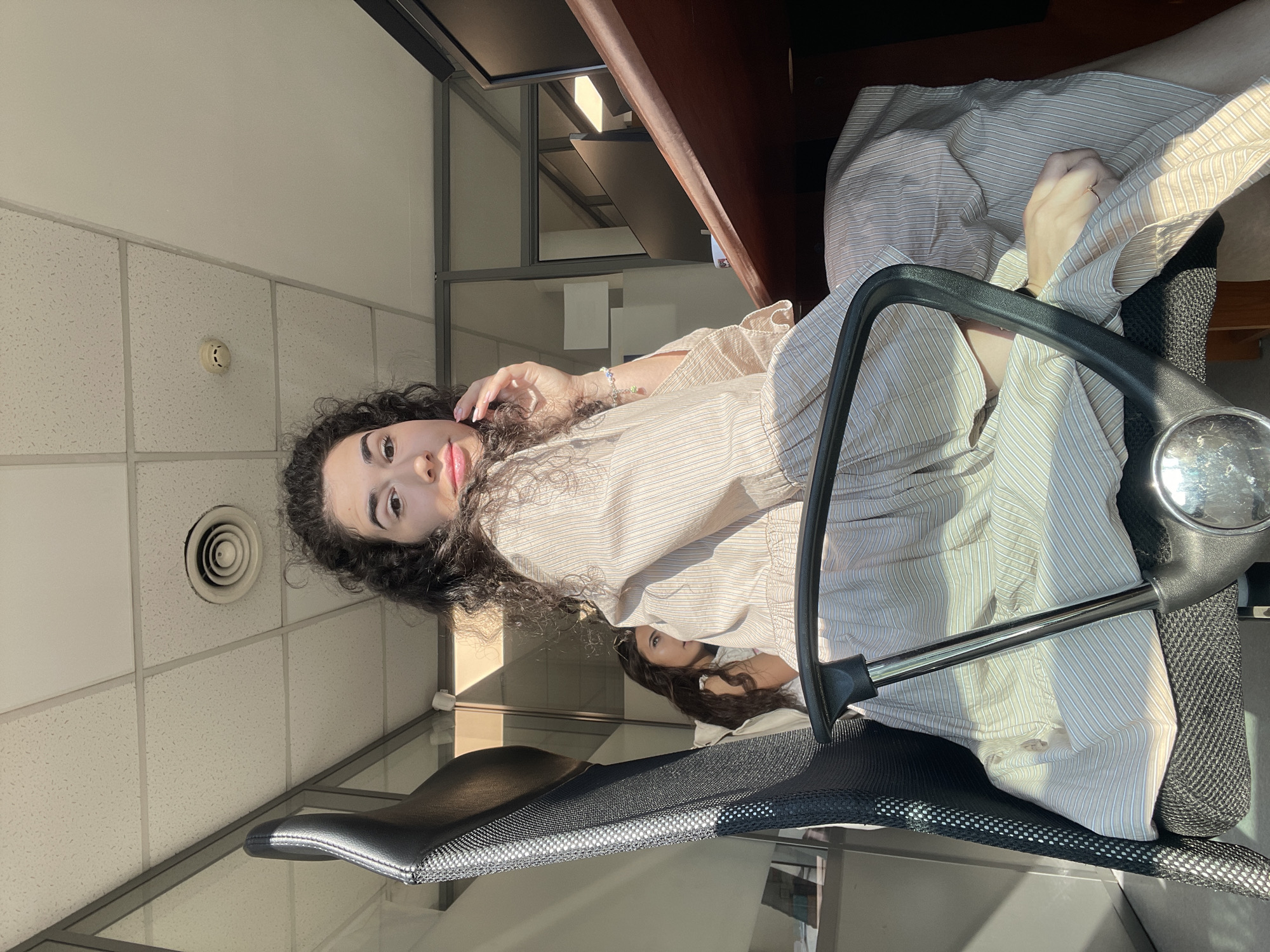 A young woman with long, dark curly hair is seated in an office chair in a sunlit office in Sofia, Bulgaria. She is wearing a light-colored, striped shirt and a matching skirt, and is looking towards the camera. Her right hand is raised to her face. Sunlight streams in from the side, casting long shadows across the room. In the background, a reflection of another woman can be seen through a glass partition. The office appears to be modern, with a suspended ceiling, fluorescent lighting, and a wooden desk with a computer monitor.