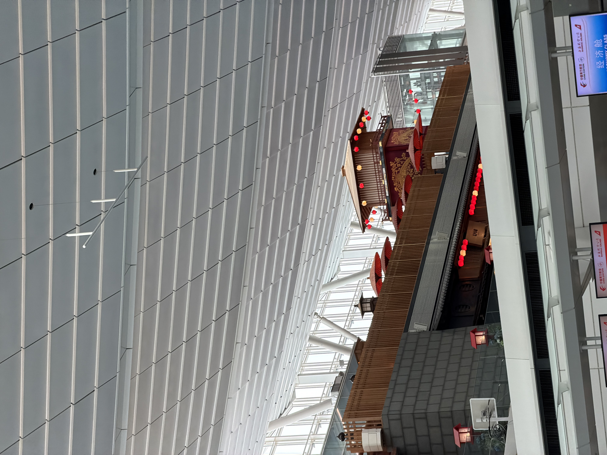 The image shows the interior of a modern building, likely an airport terminal in Ota, Japan, with a distinct Japanese-inspired architectural element. The ceiling is a complex, multi-layered structure of white, diamond-shaped panels, interspersed with light fixtures. Below the ceiling, a multi-story, wooden structure with traditional Japanese architectural features is visible. This structure is adorned with red lanterns and decorative elements, including red umbrellas. The facade of the lower levels features a dark, textured material resembling brick, topped by wooden railings and panels. Several windows and a modern glass elevator shaft are also present in the background, suggesting a public space. The overall impression is a blend of contemporary design and traditional Japanese aesthetics.