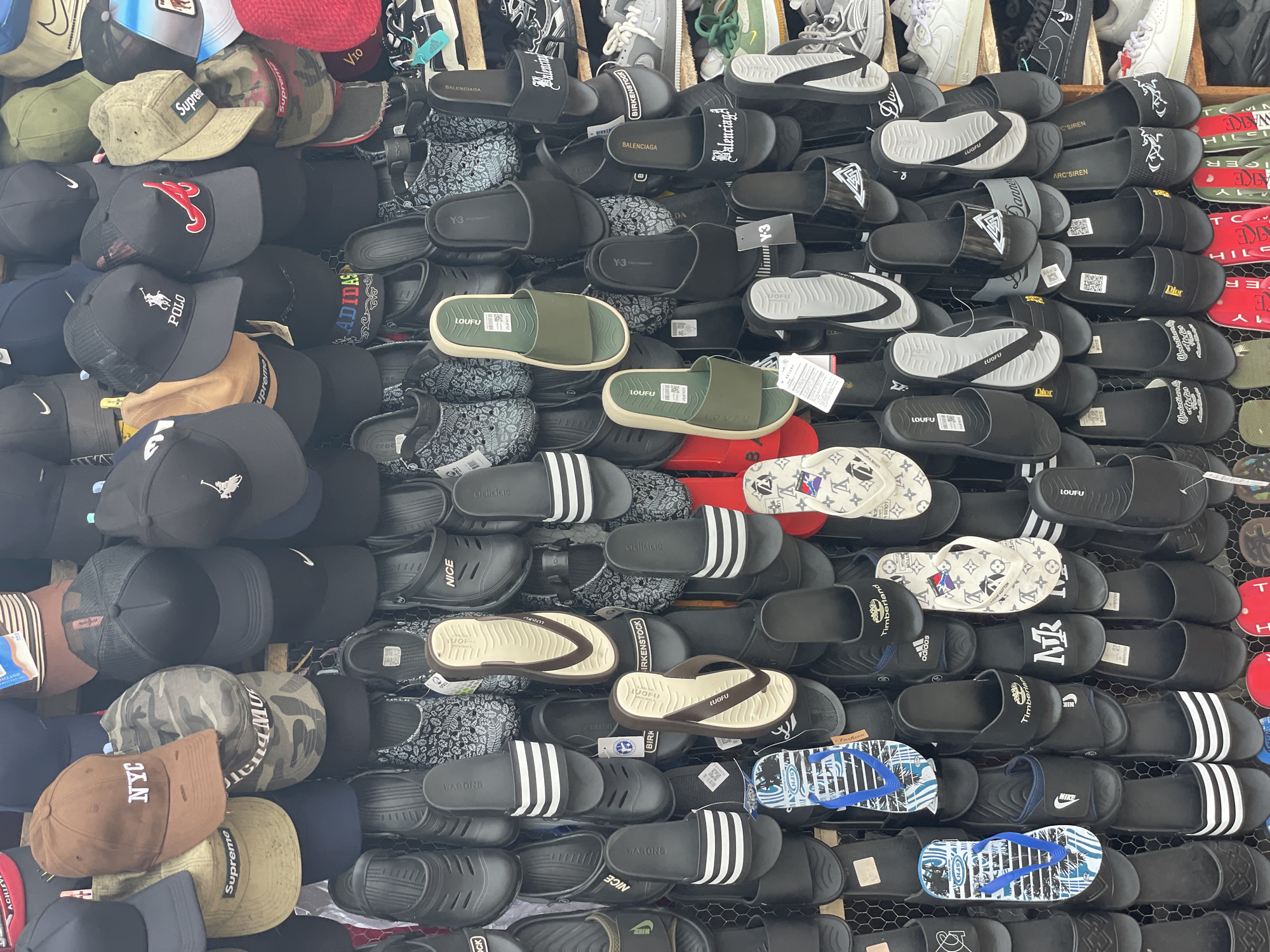 This image captures a vibrant, densely packed display of various fashion accessories, primarily footwear and headwear, likely in a market stall or a small retail shop in Badagry, Nigeria. No people are visible in the frame; the focus is entirely on the merchandise.
The upper portion of the image shows numerous baseball caps and trucker hats stacked and hung. Below, a vast assortment of sandals, slides, flip-flops, and clogs are meticulously arranged, often in rows or stacks, filling the majority of the frame. Many items have small white tags, suggesting they are for sale. The display method, with items tightly packed on what appears to be vertical racks, is typical of bustling market environments.
Notable details include a wide array of visible text and brand logos. On the headwear, brands such as "Supreme," "POLO" (resembling Ralph Lauren), "NYC," "ADIDAS," "NIKE," "Timberland," "BALENCIAGA," "Y-3," "V10," and "LOUFU" are seen. The footwear section prominently features "BALENCIAGA," "Y-3," "LOUFU," "BIRKENSTOCK" (style), "ADIDAS," "NIKE," "WAGONG," "NICE," "CROCS" (style), "TIMBERLAND," "MR," "DIOR," "ARC'SIREN," and "Danner." Some flip-flops also display patterns similar to the Louis Vuitton monogram. The mix of well-known international brands and less common names, some possibly imitations, is characteristic of street markets.
The scene depicts a commercial setting designed to showcase a large quantity of goods, offering a glimpse into the retail landscape of the area.