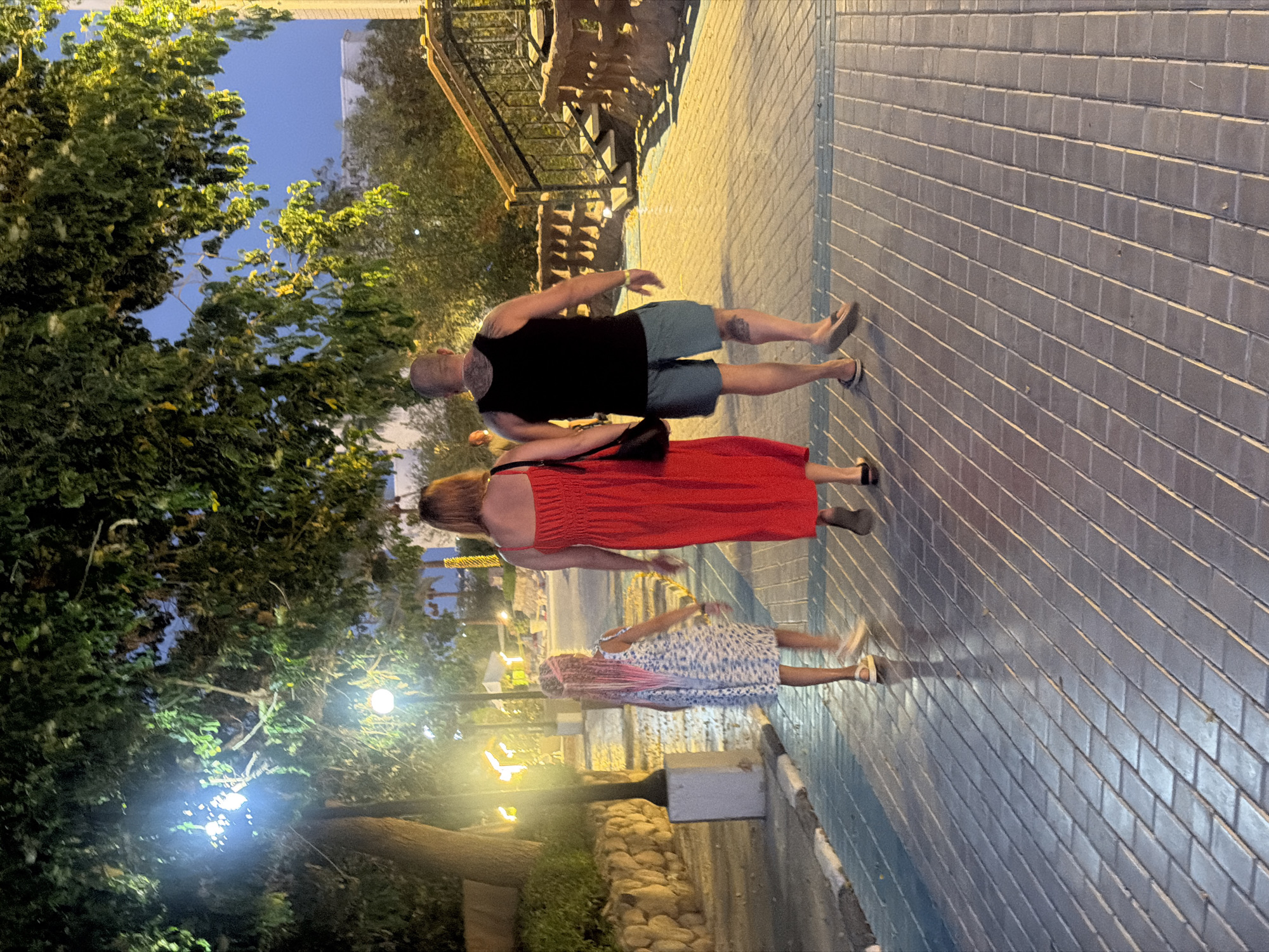 A family walks along a tiled pathway in Sharm El Sheikh, Egypt. A man, a woman, and a young girl are walking away from the camera, heading towards a set of stairs with a railing. The man is wearing a black tank top and blue shorts. The woman is wearing a red dress and carrying a black purse. The girl has long pink braided hair and is wearing a white and blue floral dress. The pathway is illuminated by overhead lights and streetlights, casting shadows on the ground. The environment appears to be a resort or a public area with greenery and stone walls in the background.