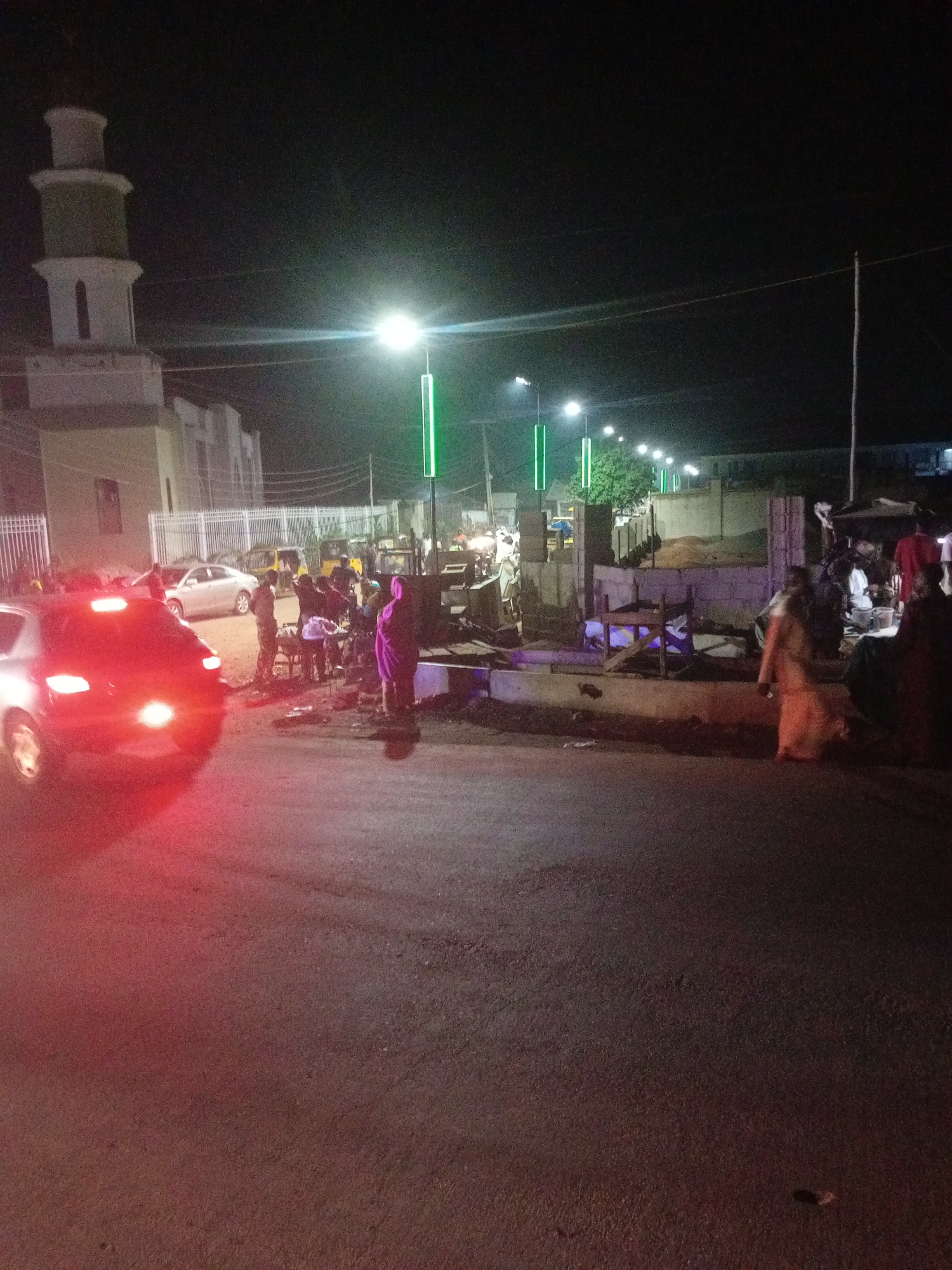 In Tundun Wada West, Nigeria, at night, a street scene unfolds with a prominent mosque minaret on the left. Several cars, including a red one with illuminated taillights in the foreground, are present. Groups of people are gathered on the side of the road, some standing near stalls or makeshift structures. Streetlights illuminate the scene, with some featuring green lighting elements. The ground appears to be asphalt, with debris scattered in some areas. The overall impression is one of a bustling evening in a commercial or gathering area.