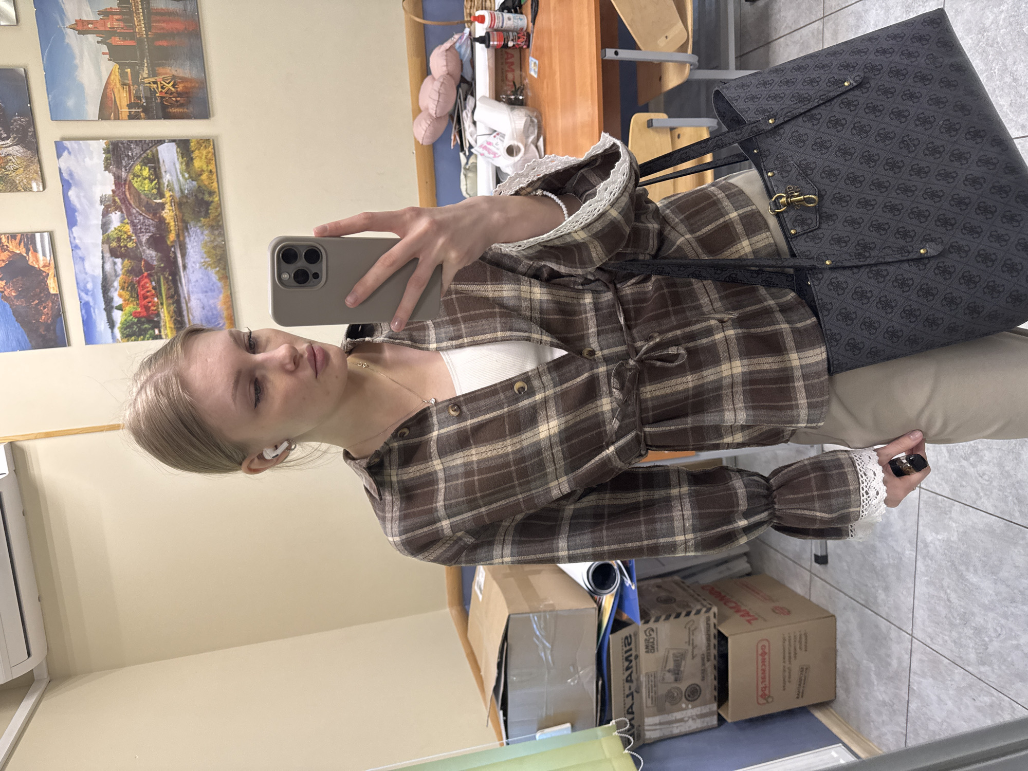 A young woman with blonde hair is taking a selfie in what appears to be an interior space. She is wearing a brown and tan plaid shirt with white lace trim on the sleeves and collar, and a white top underneath. She is holding a cell phone with a gray case up to her face to capture the image. Her left ear has a white wireless earbud in it.
Behind her, there are various elements suggesting a functional space, possibly an office or classroom. There's a desk with items on it, including bottles and other small objects. Stacked boxes and a blue bin are visible near the floor. On the wall, several pictures or paintings of landscapes, including one that appears to be of the city of Perm, Russia, are hung. The overall environment is well-lit and appears to be indoors. A large gray patterned tote bag is hanging near the young woman.