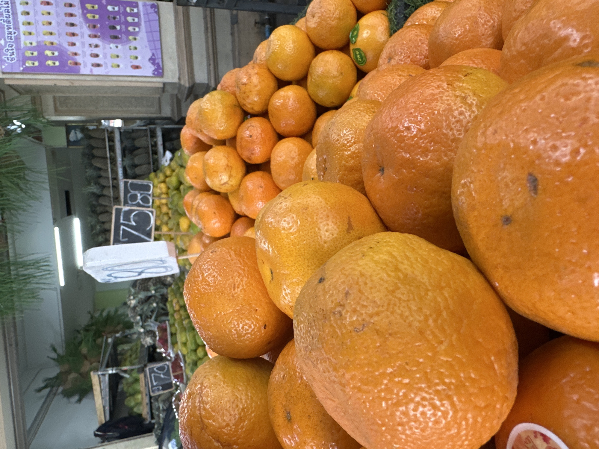 The image depicts a close-up view of a fruit stall in Phuket City Municipality, Thailand. The foreground is dominated by a large pile of bright orange tangerines, their textured skins catching the light. Behind the tangerines, more fruit, possibly limes or small green apples, are visible, arranged on shelves. In the background, a sign with the numbers "75 80" is propped up, likely indicating prices. Further back, rows of pineapples are displayed on shelves, suggesting a diverse fruit market. A purple poster with images of various drinks hangs on a pillar to the right. The lighting suggests an indoor or covered market setting.