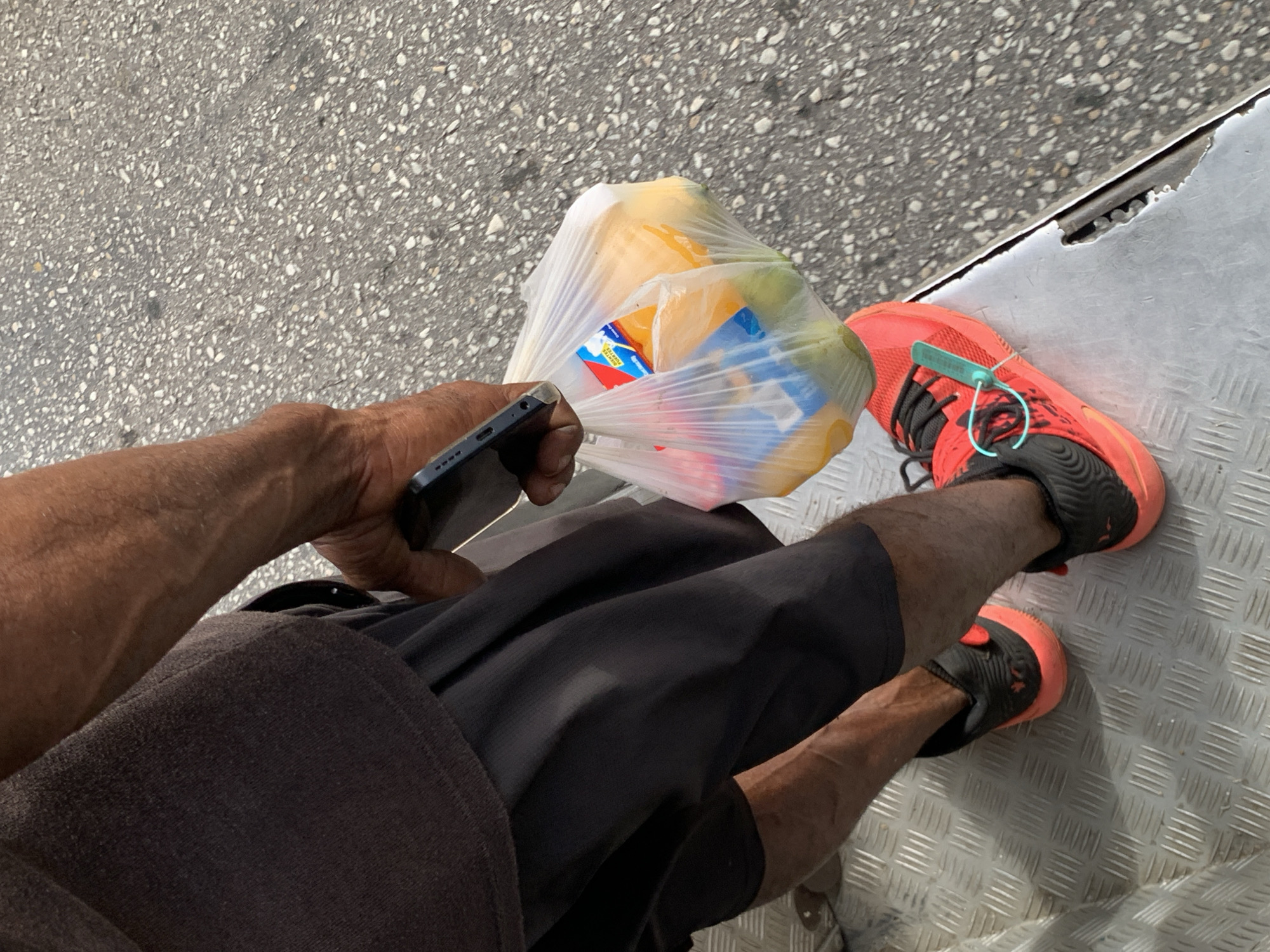 An overhead, high-angle shot captures a person's lower body, hands, and feet on a bright day. The individual, with dark skin, is dressed in dark shorts or pants, with a dark fabric draped over their left leg. They are wearing black and orange athletic shoes with black laces, and a light blue plastic security tag or zip tie is visible on the laces of the right shoe.
In their left hand, the person holds a dark-colored smartphone, while their right hand grips a clear plastic shopping bag. Inside the bag are several yellow items, likely fruits such as mangoes or papayas, and a small box or package with blue and red markings, where the word "POCKET" is partially visible.
The ground surface is divided; on the left, there is grey asphalt with small white aggregate, and on the right, a textured metallic surface (diamond plate) extends. A clear seam marks the transition between these two surfaces. The bright lighting suggests it is daytime, possibly sunny. The scene depicts a casual moment, perhaps someone waiting or moving between locations, carrying groceries and using a phone. The location is specified as La Asuncion, Venezuela, though no direct visual cues confirm this within the image.